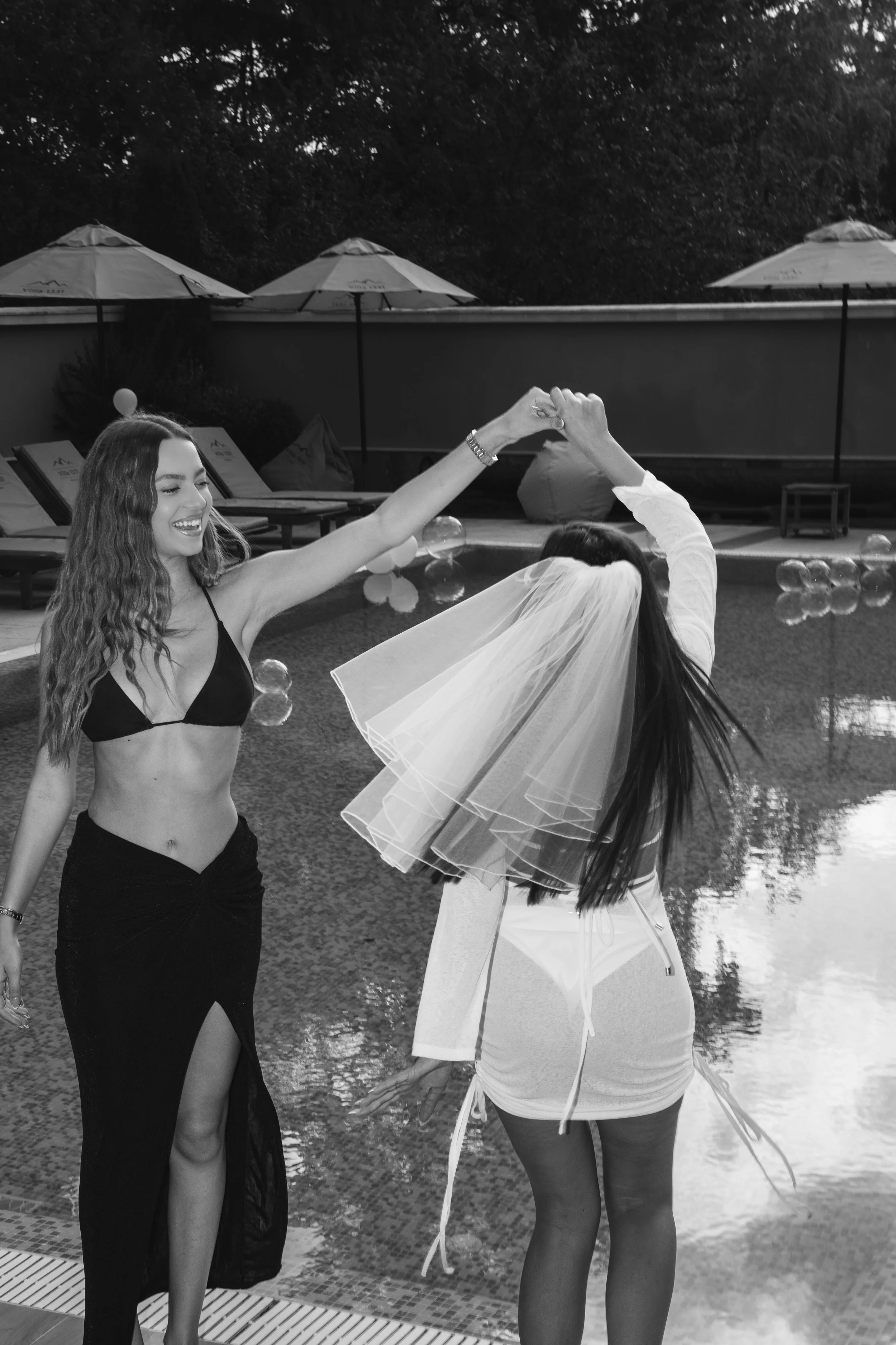 Two women dancing by a poolside, one in a bikini top and flowing skirt, the other in a dress with a veil, under poolside umbrellas, in black and white.