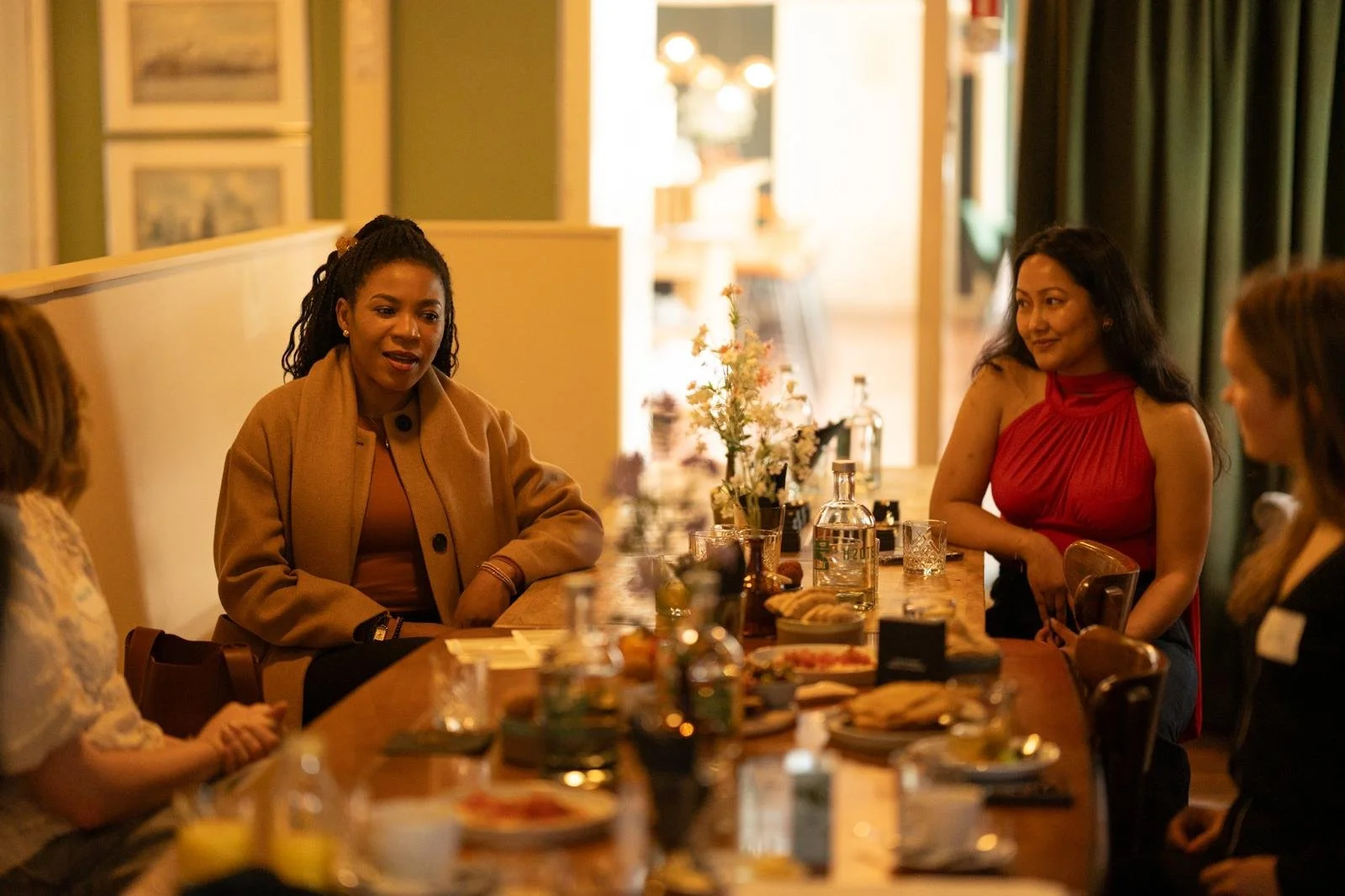 Four women sitting around a wooden table with food, drinks, and flowers, engaged in conversation in a warmly lit restaurant or cafe.