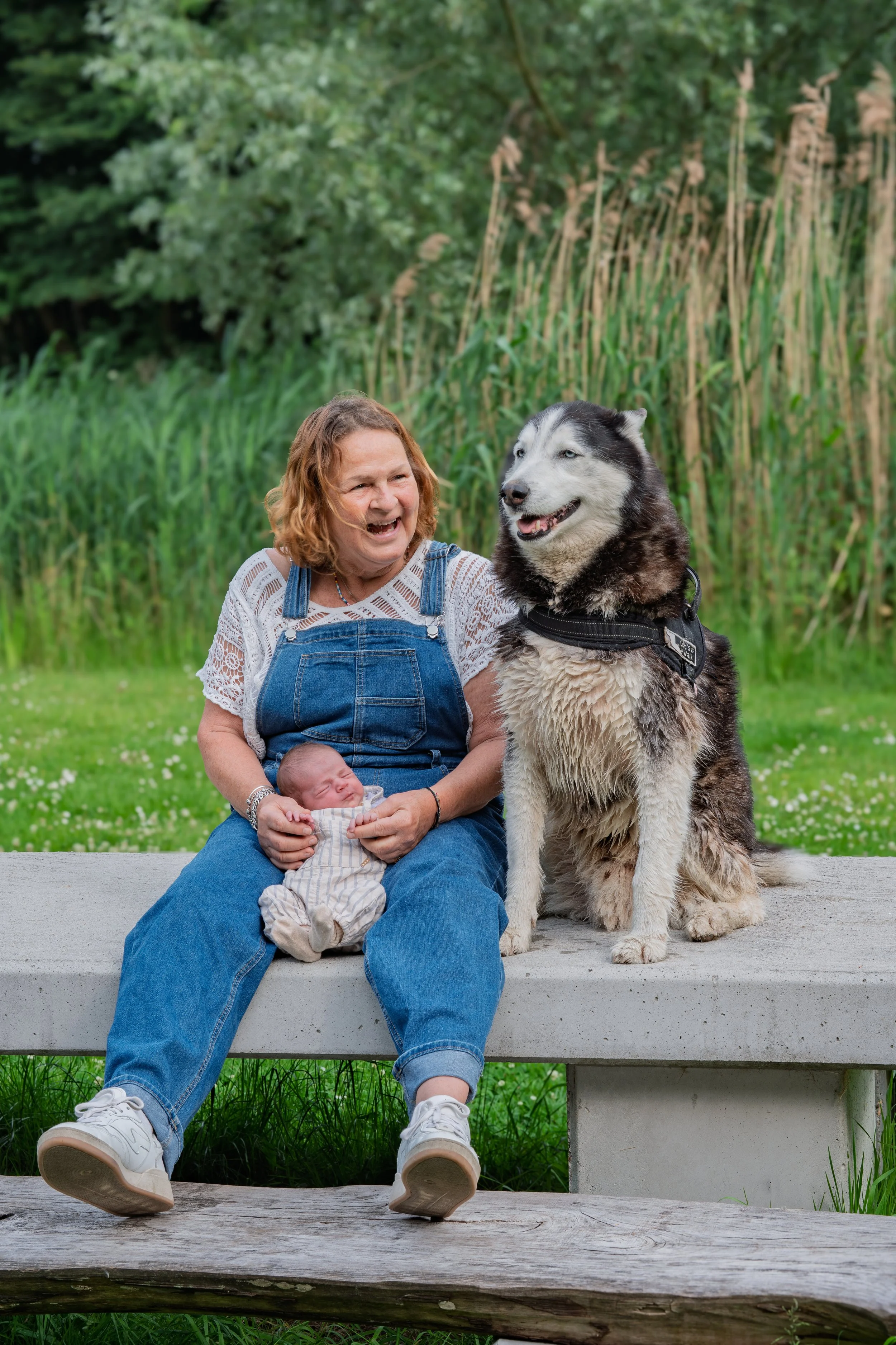 A woman with curly hair wearing a white crochet top and denim overalls sitting on a concrete bench outdoors. She is holding a newborn baby dressed in light-colored clothes with stripes, and there is a large Siberian Husky dog sitting next to her. The