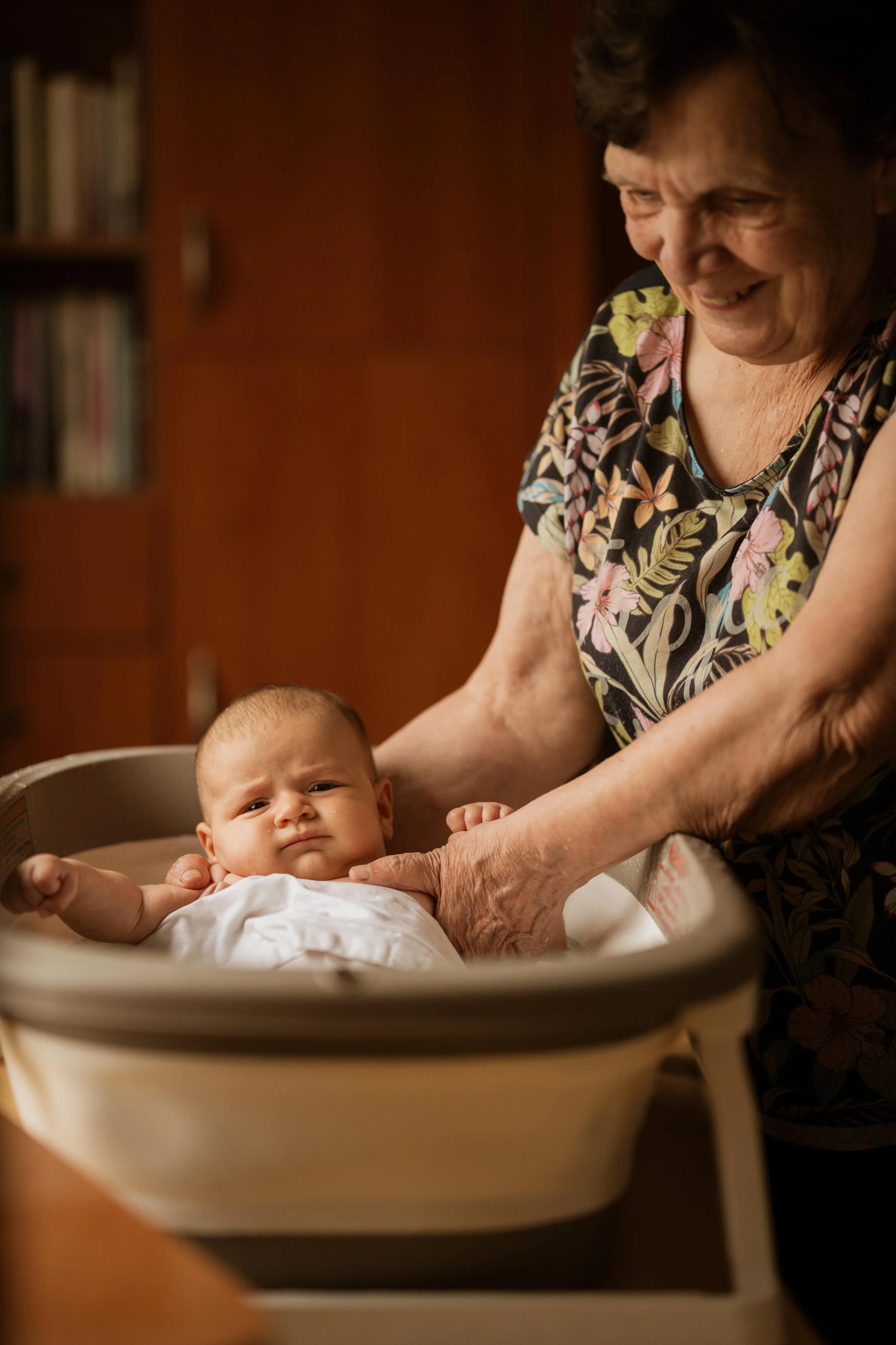 An elderly woman holding a baby in a bassinet, smiling and looking at the baby.