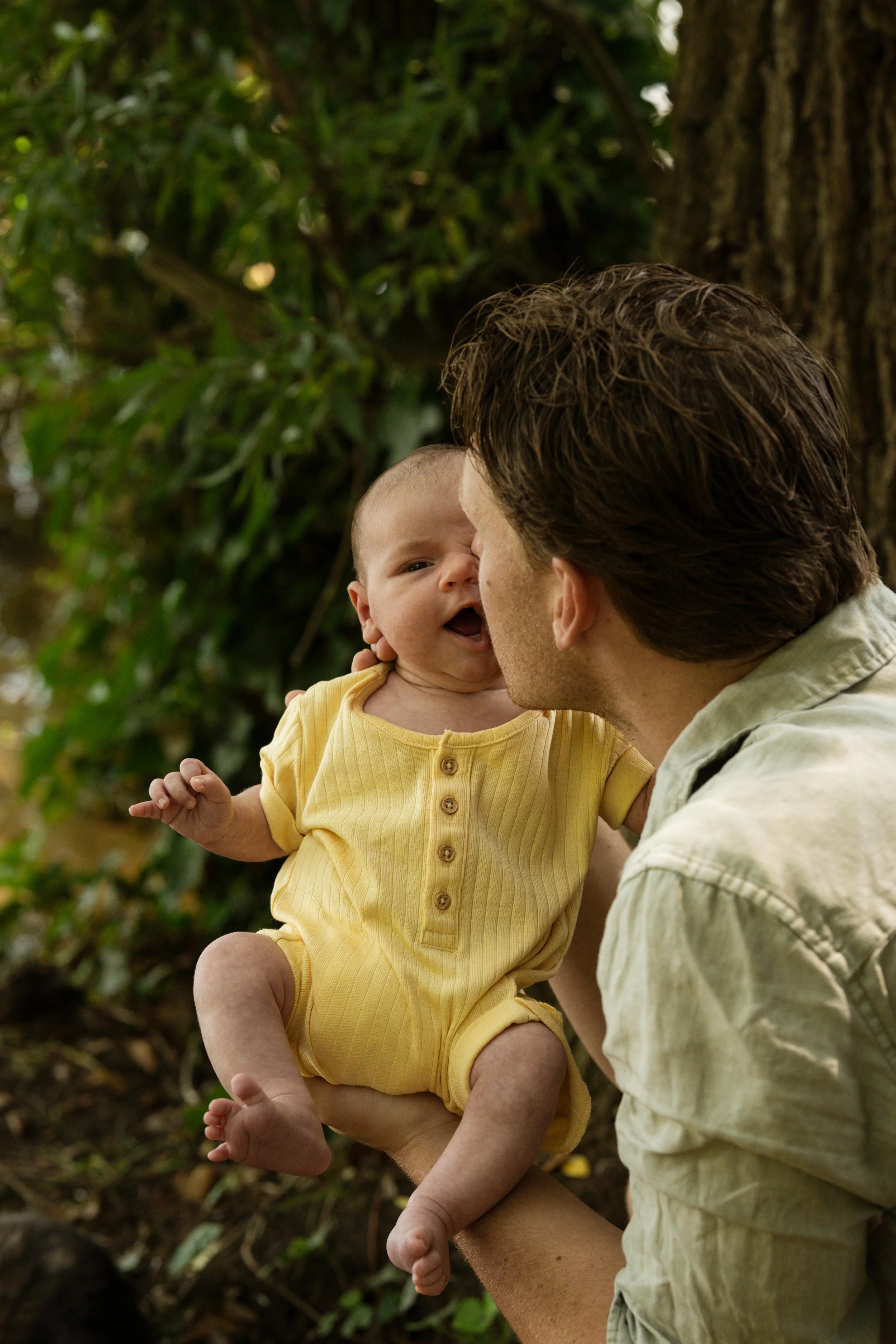 A man holding a baby outdoors near a tree, with both making a playful face.