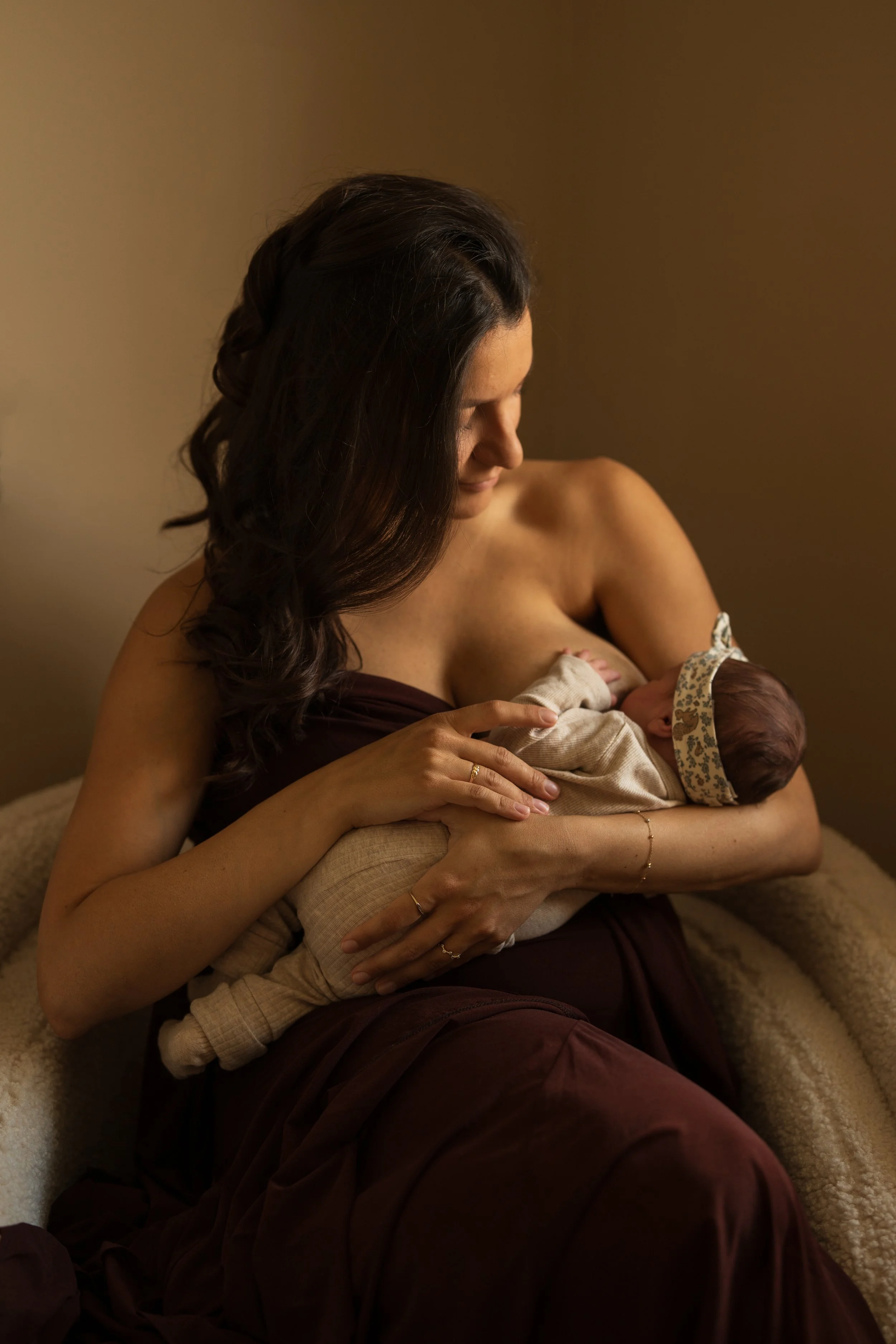 A woman with dark hair, wearing a strapless dress, breastfeeding a baby with a floral headband, seated on a soft chair in warm lighting.
