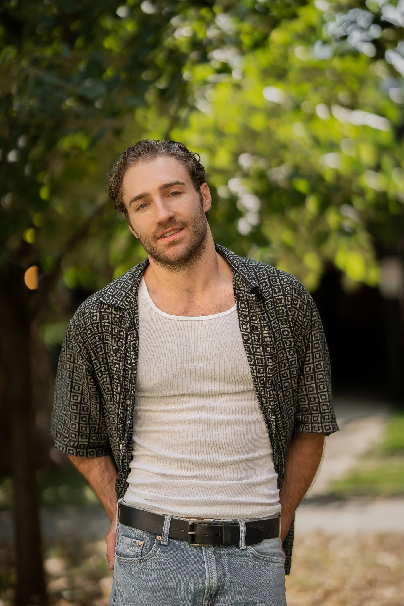 A young man with brown hair and a beard, wearing a patterned short-sleeve shirt over a white t-shirt, standing outdoors with green trees in the background.