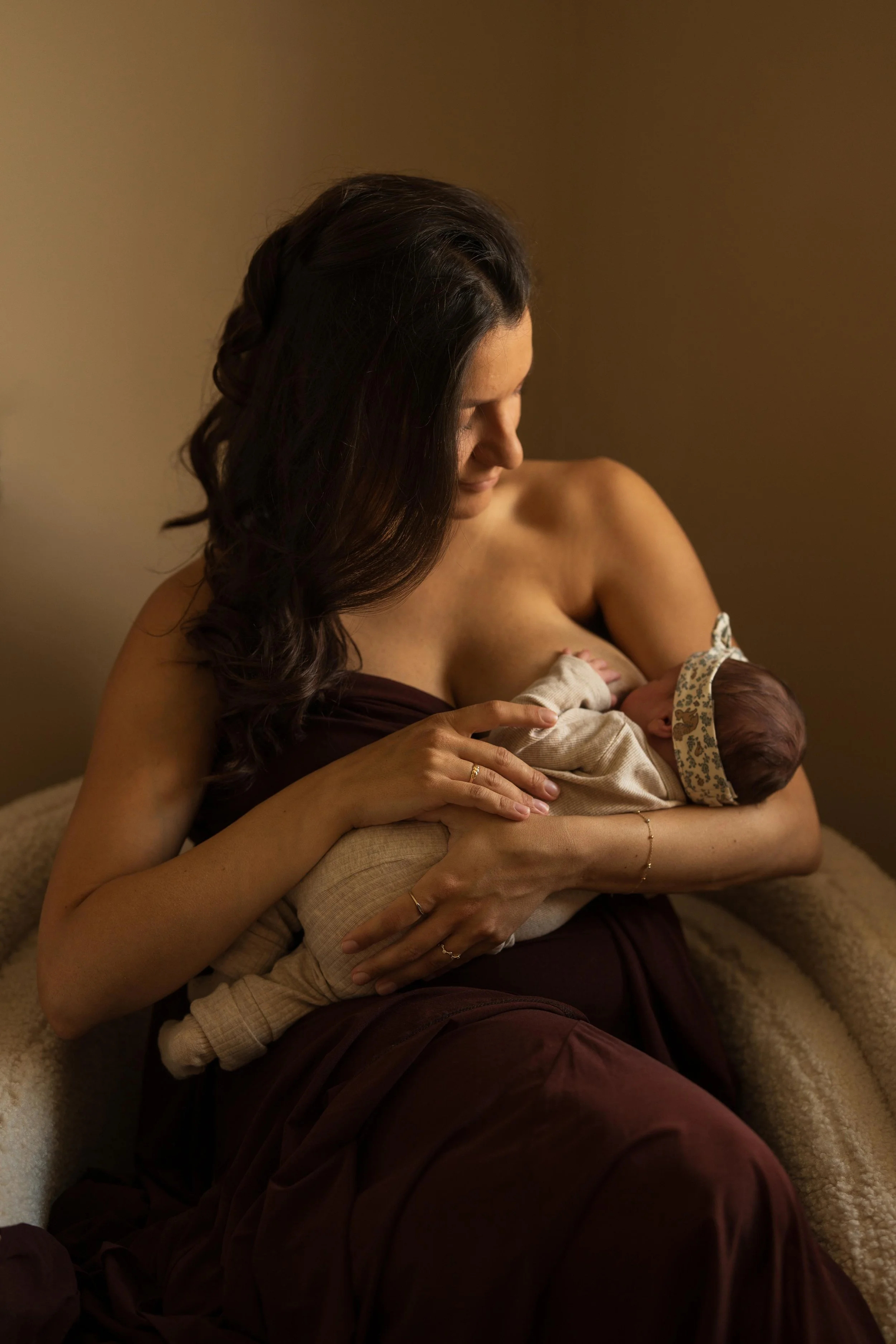 A woman with dark brown hair breastfeeding a newborn baby with a headband, in a warm, softly lit room.