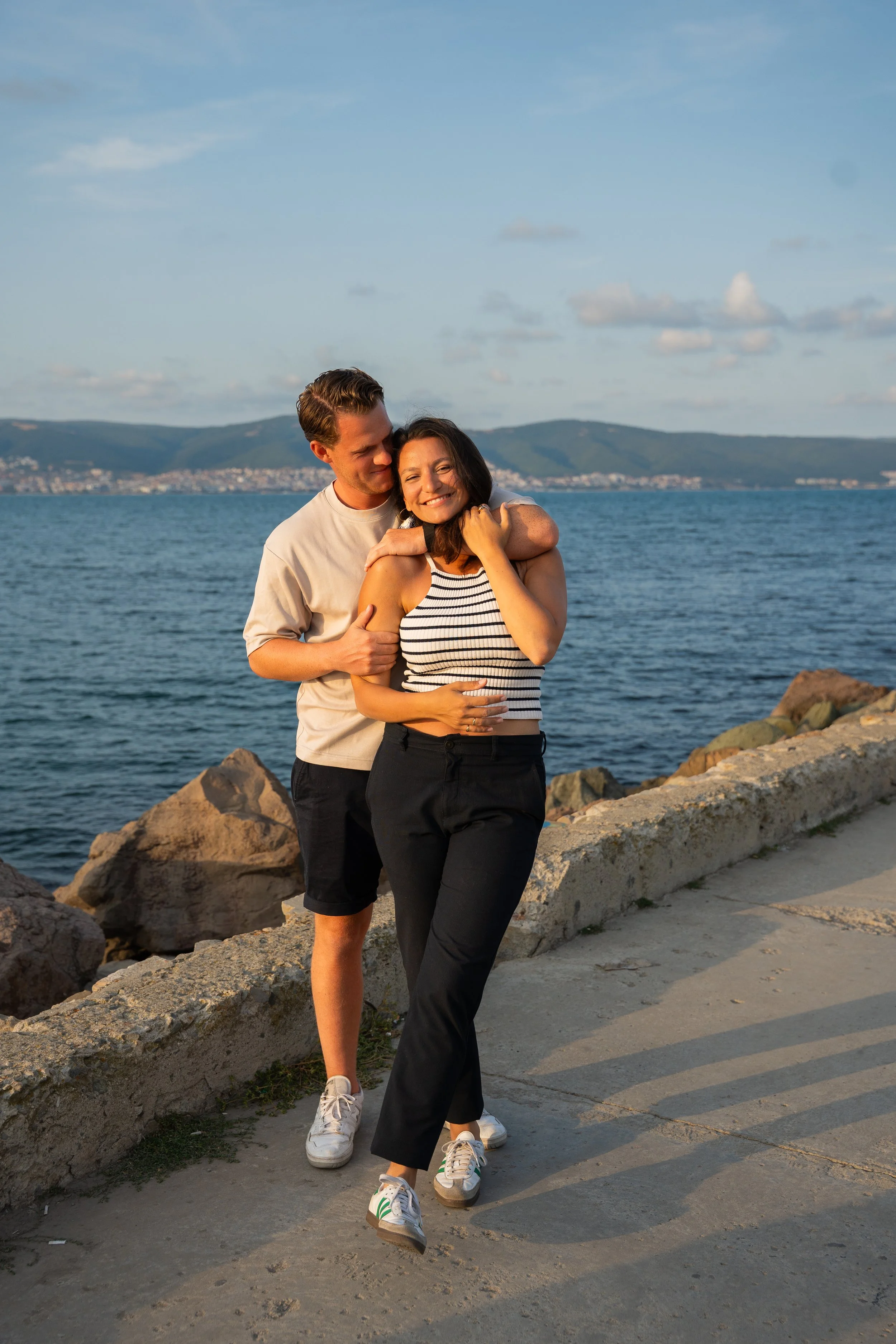 A young couple is walking along a waterfront, with the man embracing the woman from behind, both smiling, during sunset. They are near rocks and a concrete path, with water and mountains in the background.