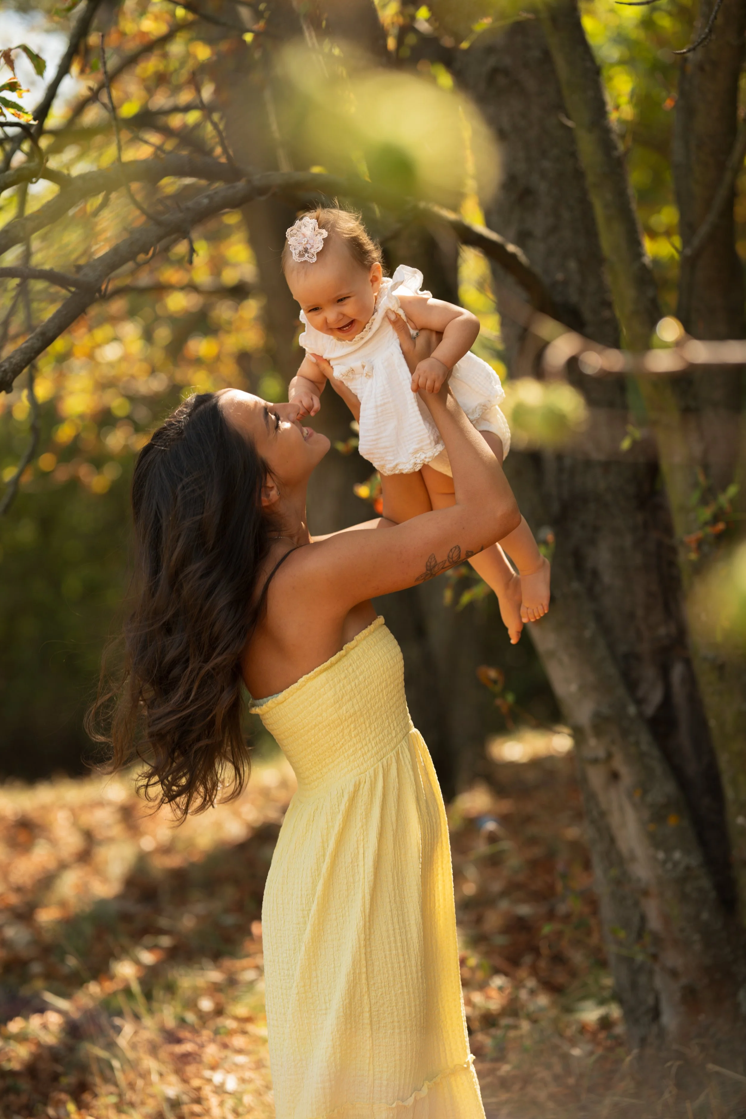 A woman in a yellow dress lifting a smiling toddler girl in a white dress with a flower headband, outdoors among trees with autumn leaves.