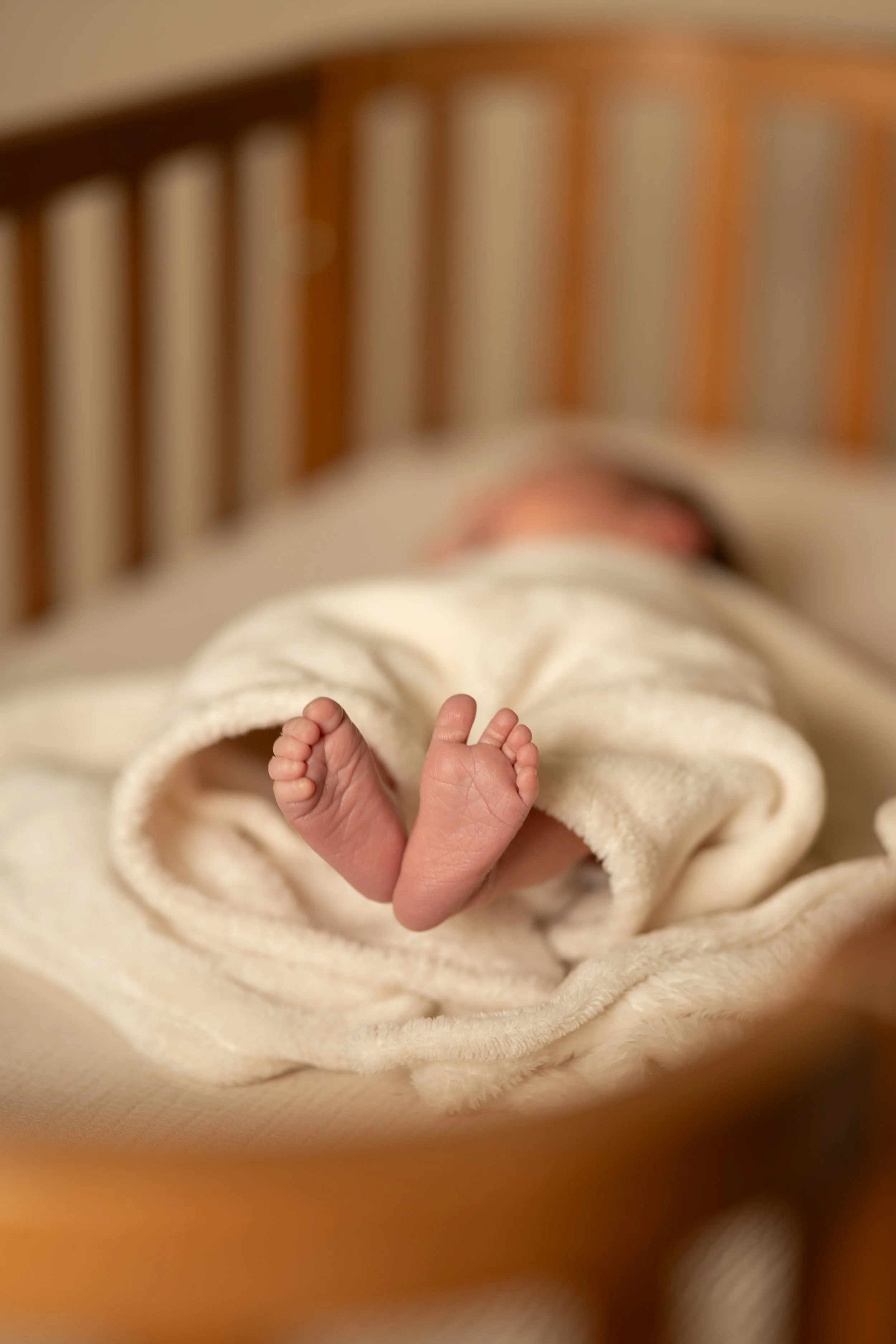 A newborn baby's feet poking out from under a soft, cream-colored blanket on a bed with a wooden headboard.