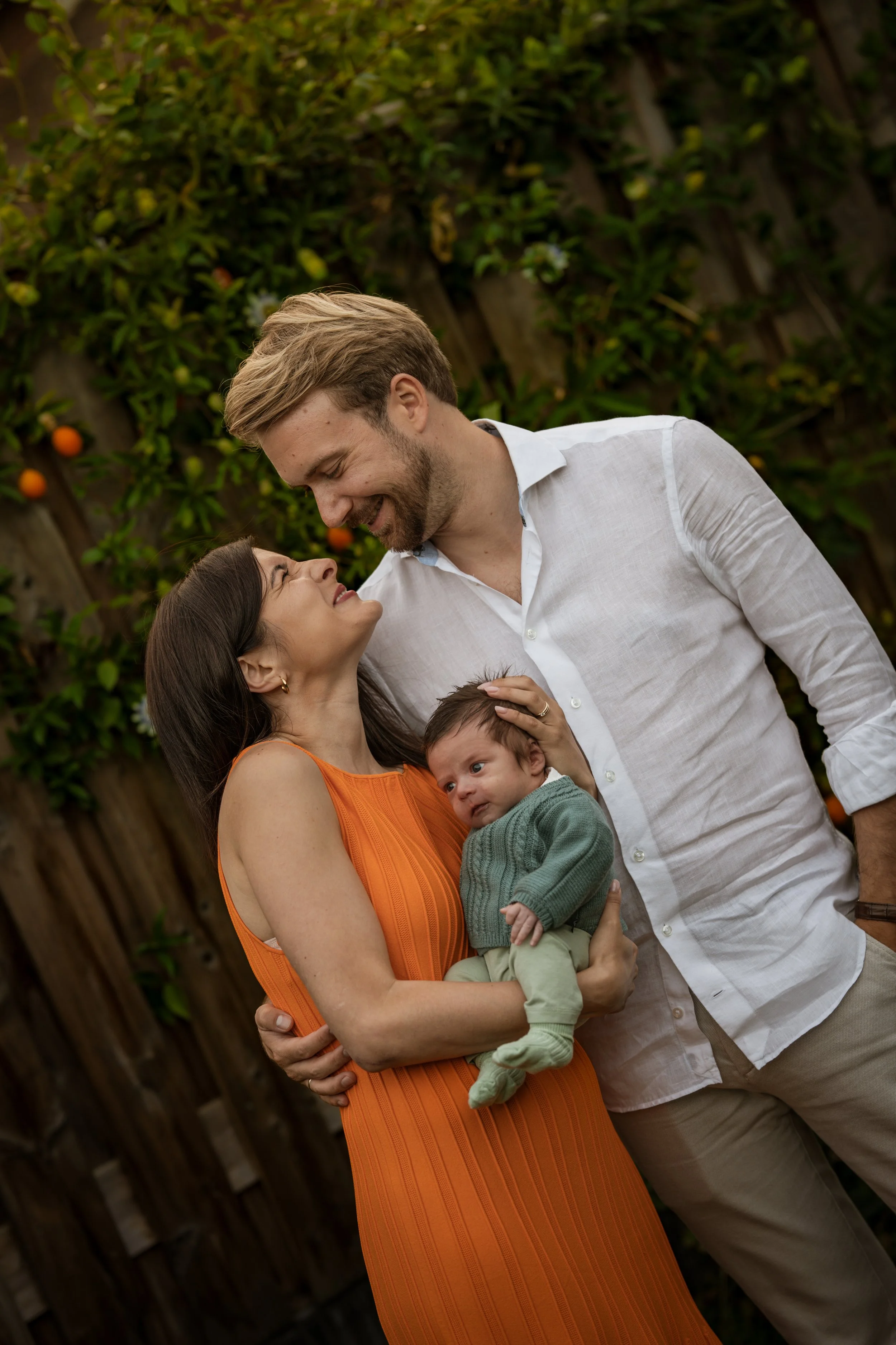 Family of three smiling and looking at each other outdoors, with a wooden fence and greenery with orange fruits in the background. The woman wears an orange dress, the man a white shirt, and the baby green clothes. Newborn photo shoot, warm, outdoor,