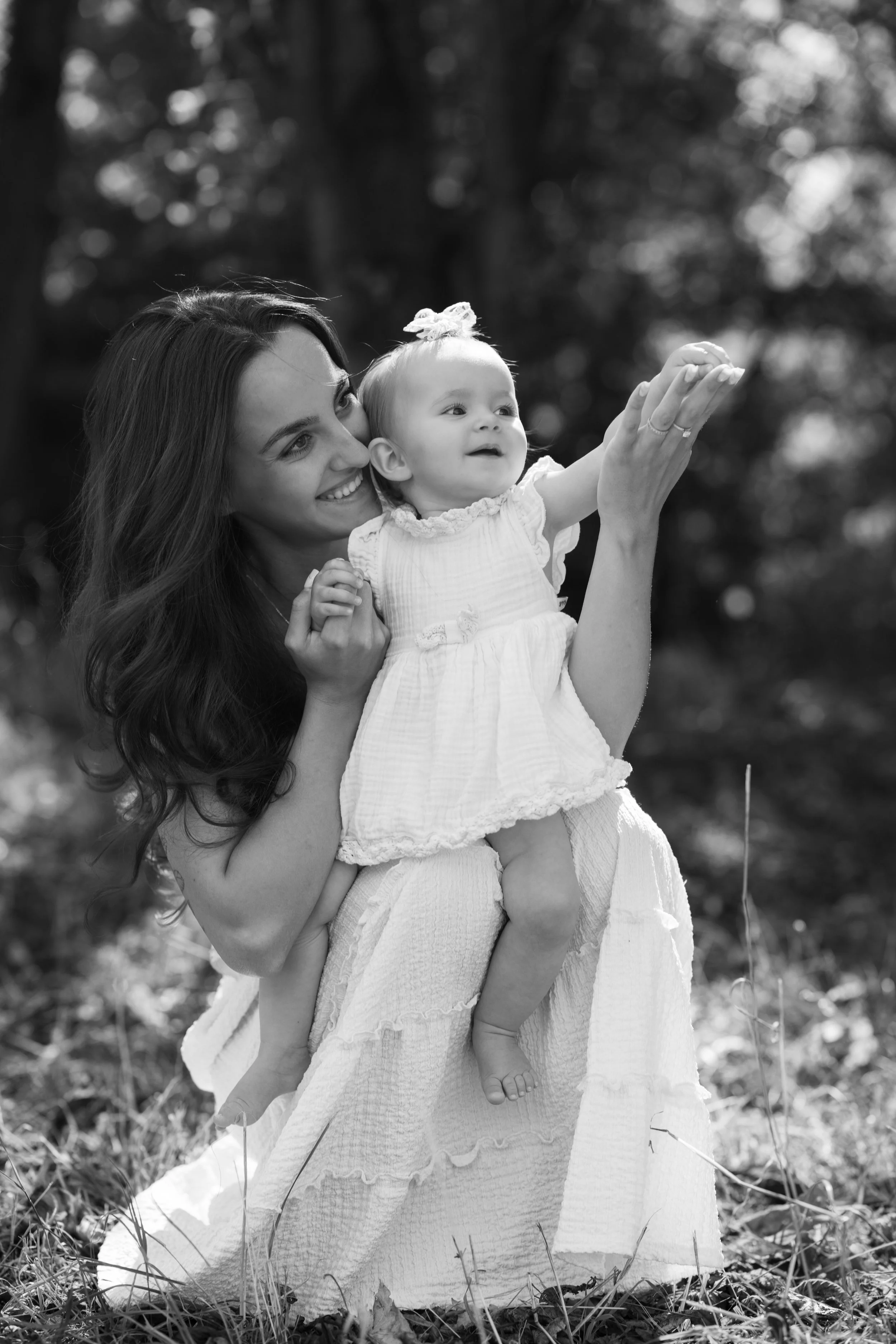 A woman holding a young girl outdoors, both smiling, as the girl reaches out with her hand, in a natural setting with trees in the background.