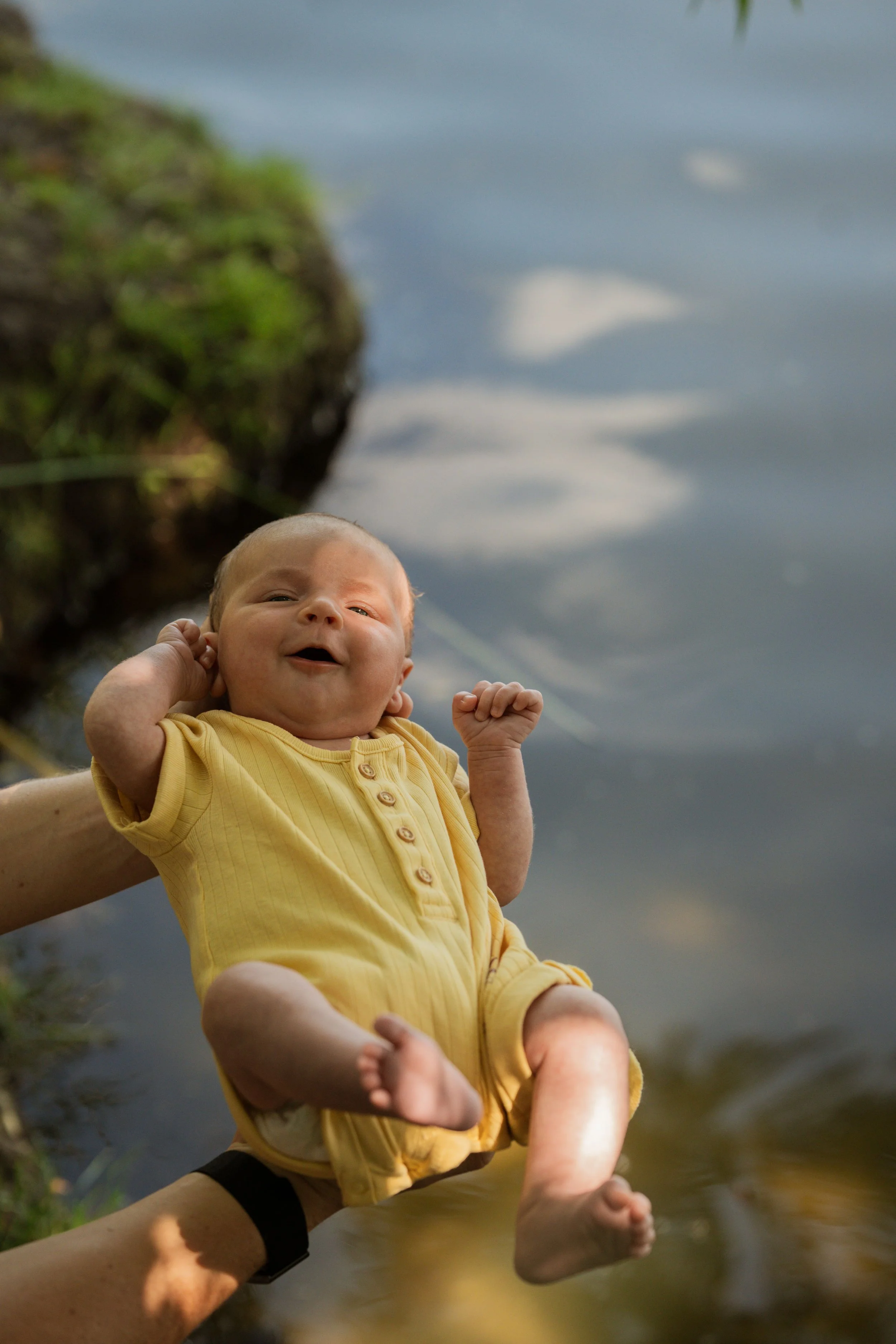 A smiling baby wearing a yellow outfit is being held over a body of water with a grassy bank visible in the background.