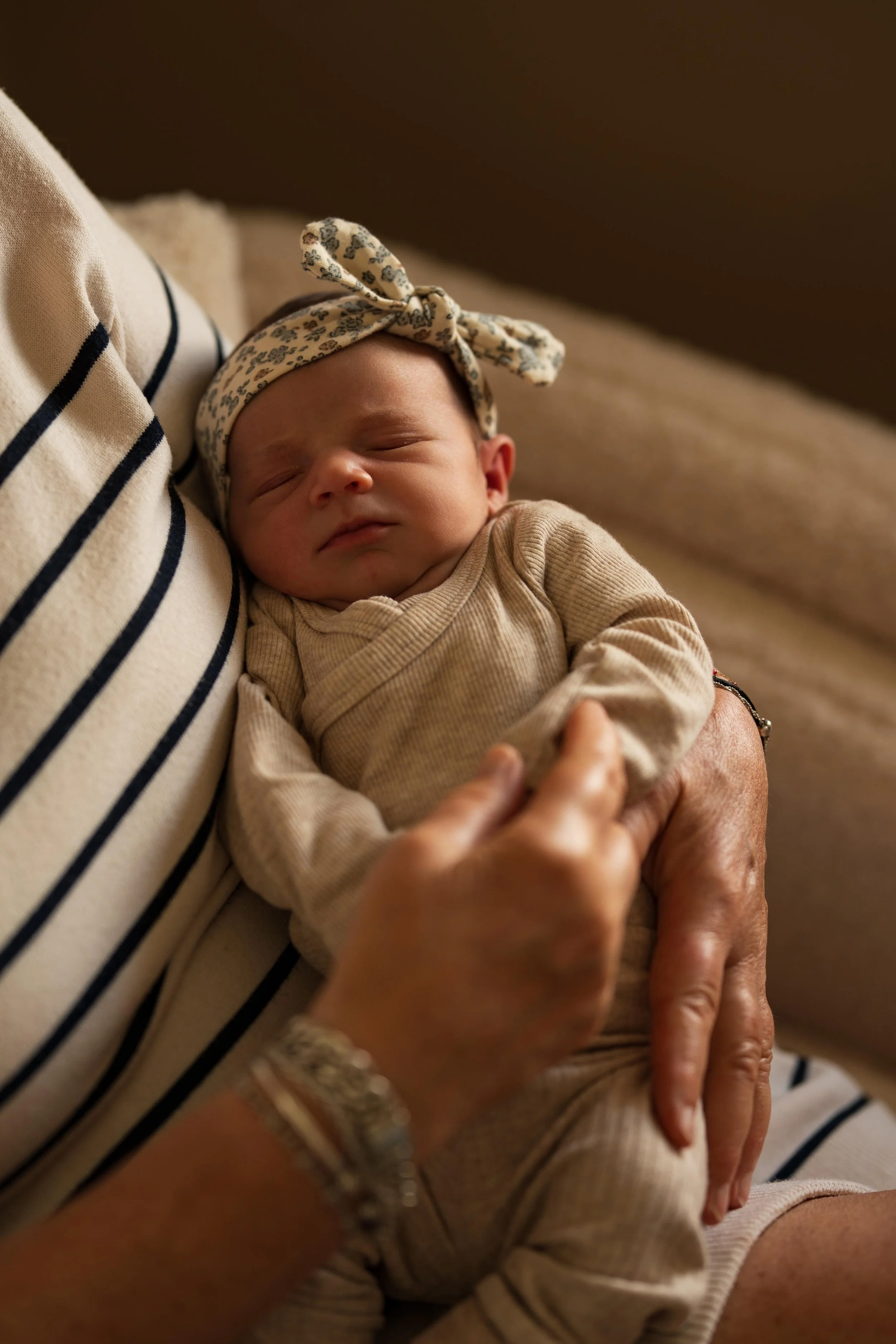 A close-up of a sleeping baby with a patterned headband, held gently in an adult's arms, wearing beige clothing.