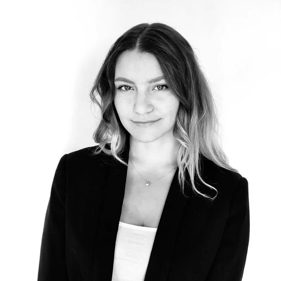 A woman with wavy hair wearing a blazer and a necklace, smiling slightly, in a black-and-white photo.