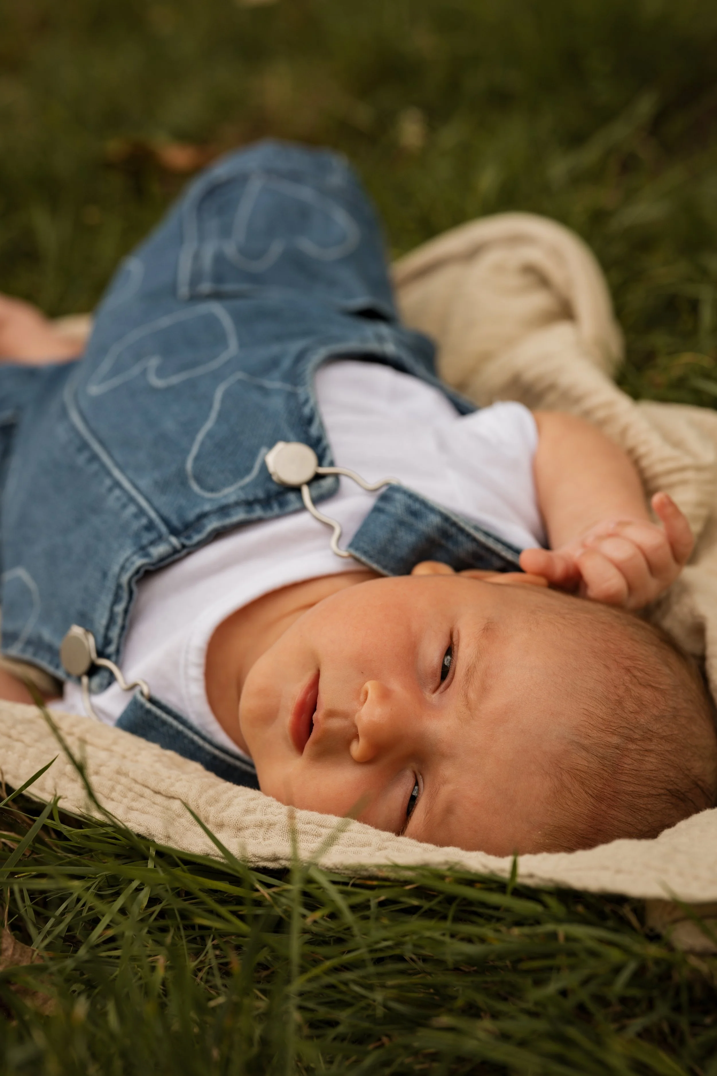 A young child lying on a blanket in the grass, wearing denim overalls and a white shirt, looking relaxed and content.