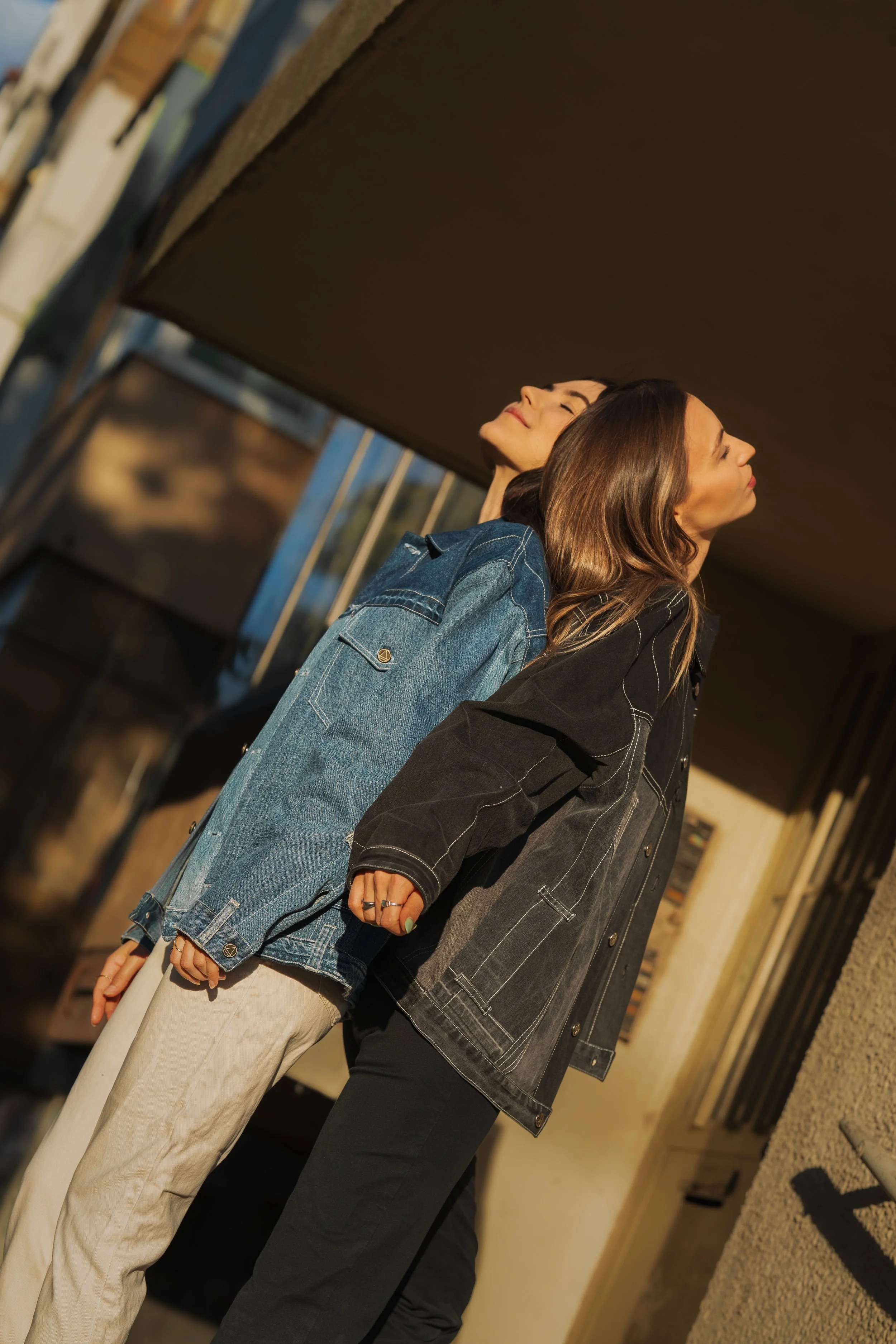 Two women standing back to back with their heads tilted upwards, both wearing denim jackets, standing outside near a building with warm sunlight.