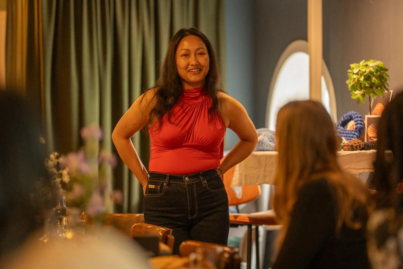 Woman in red top and black jeans standing and smiling in a cozy indoor setting with people sitting at tables, green curtains, and decorative items in the background.
