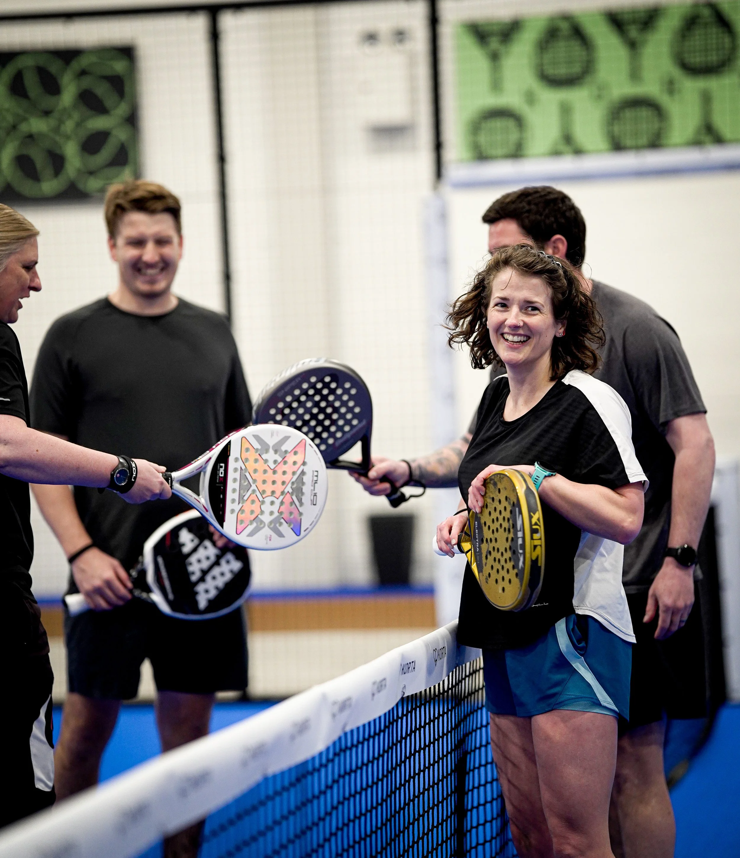 Group of four people playing padel indoors, holding padels and standing beside a net.