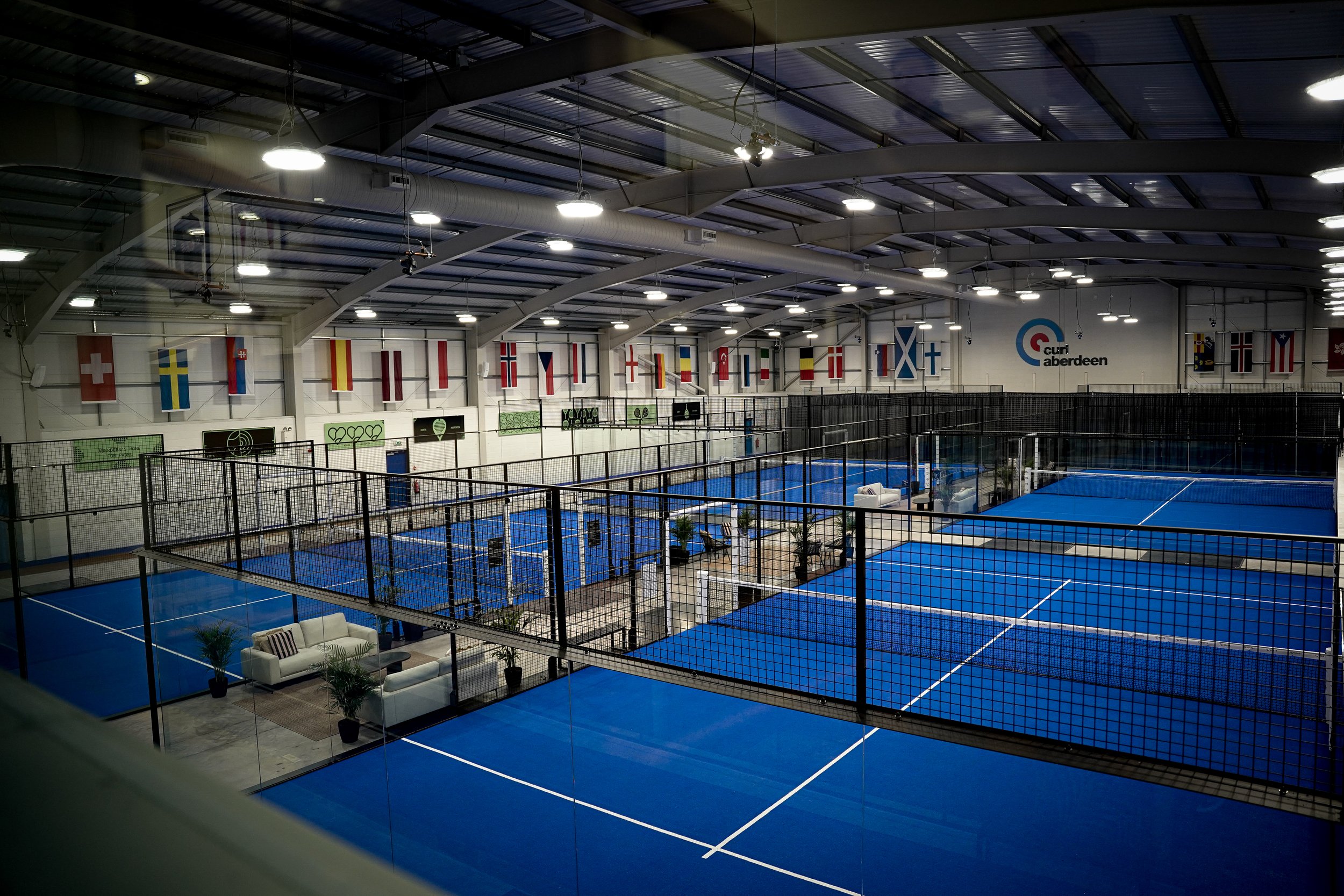 Indoor sports facility with blue courts, fencing, and flags hanging from the ceiling.
