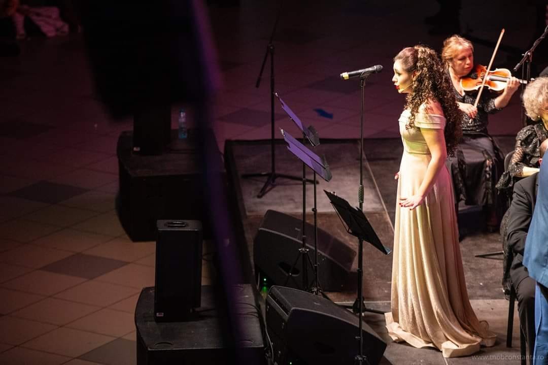 A woman with long, curly hair wearing a long, light-colored dress standing on a stage with a microphone, accompanied by musicians playing string instruments.