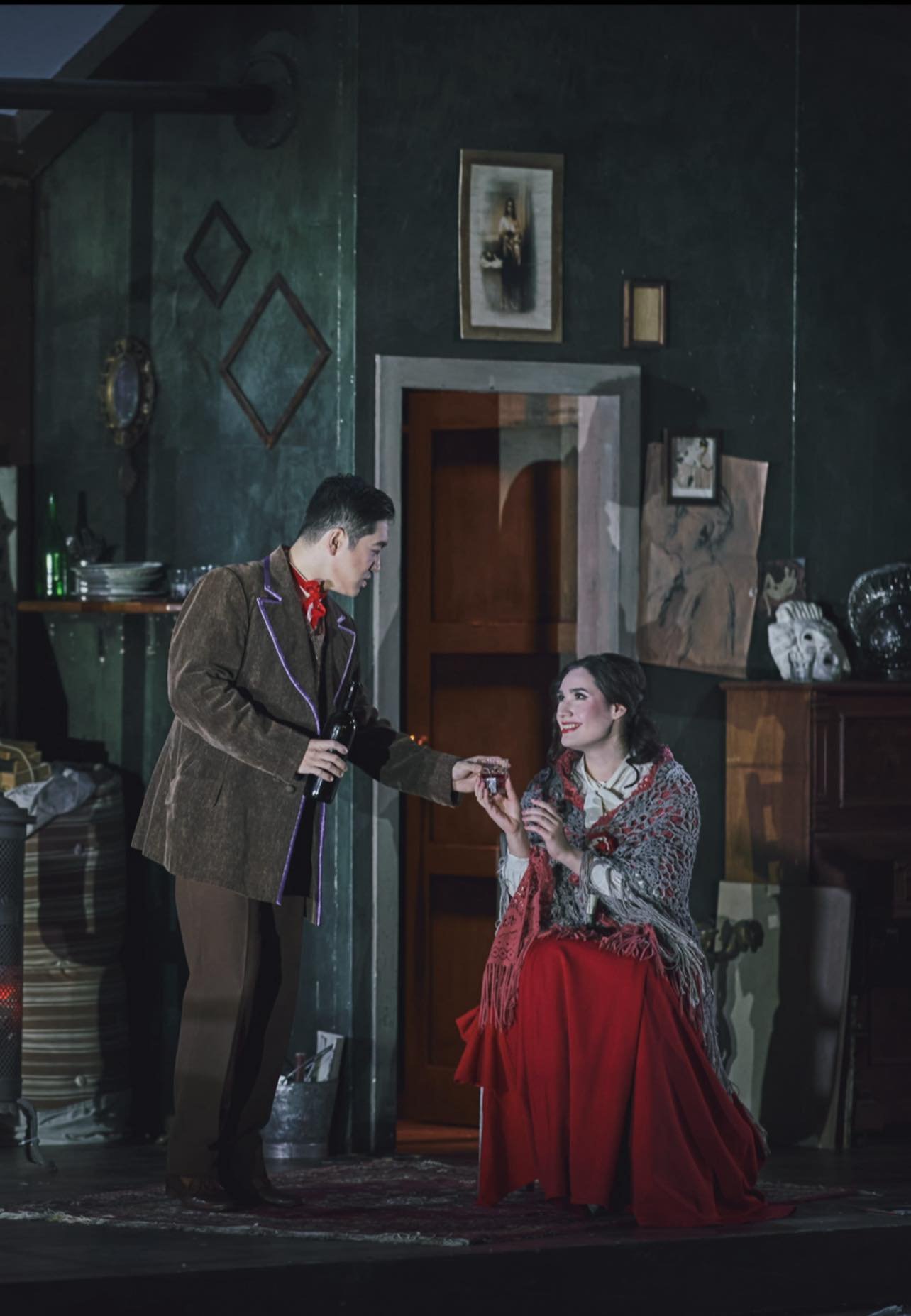 A man in a brown jacket handing a small gift box to a woman with dark hair, wearing a red skirt and a patterned scarf, in an indoor setting with dark green walls and framed artwork.