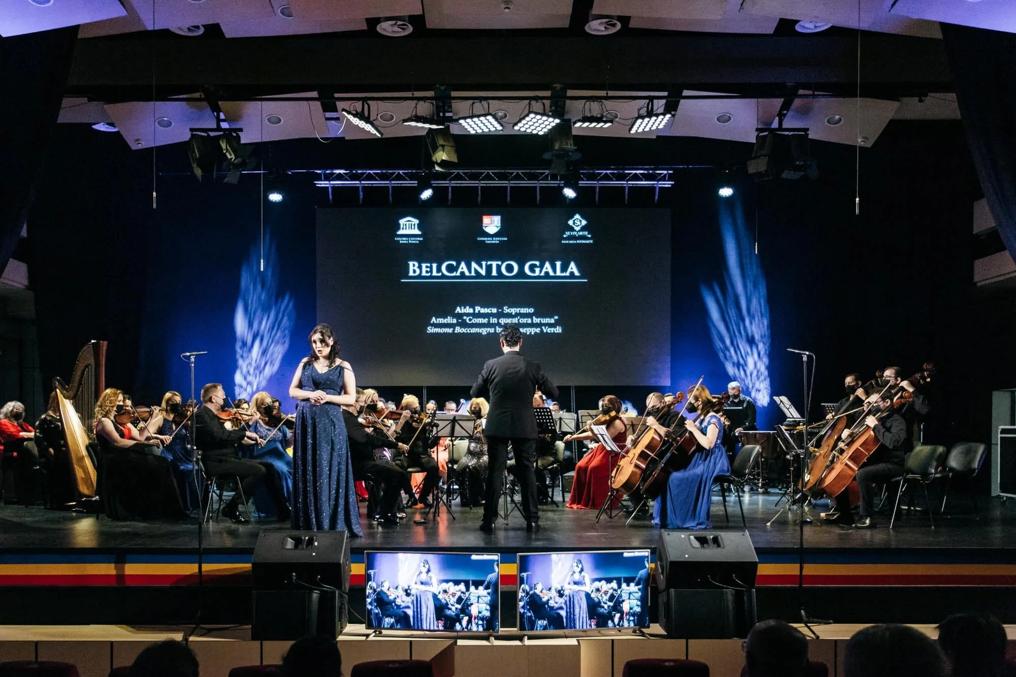 Symphony orchestra performing on stage with a woman singer in a blue gown at the Belcanto Gala, with a conductor, large screen behind displaying event details, stage lights, and screens showing the performance.