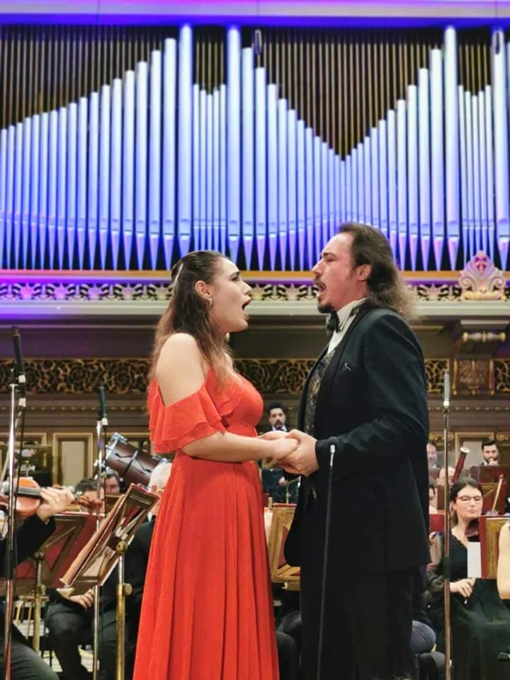 A woman in a red dress and a man in a tuxedo are singing and holding hands in a concert hall with an orchestra and a pipe organ in the background.