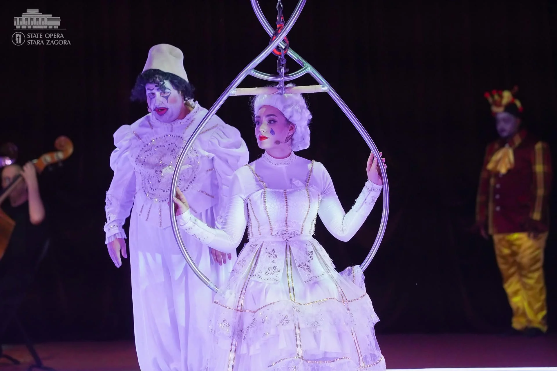 Clowns performing on stage, with two clowns in focus. The clown on the left has white face makeup, black curly hair, a white hat, and wears a white costume with elaborate embroidery. The clown on the right, dressed in a white dress with gold accents,