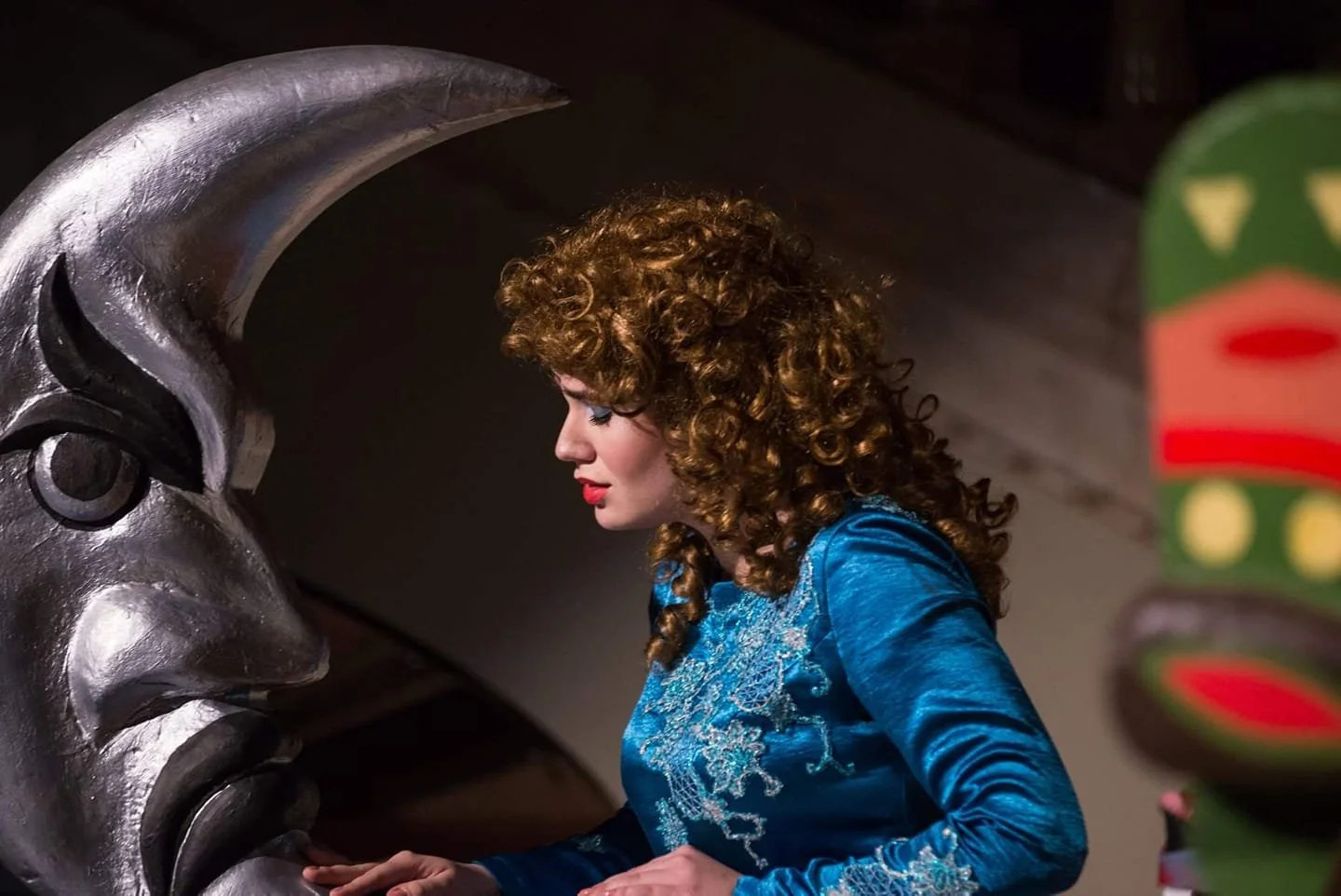 Woman with curly hair wearing a blue dress looking at a large metallic sculpture of a face with closed eyes.