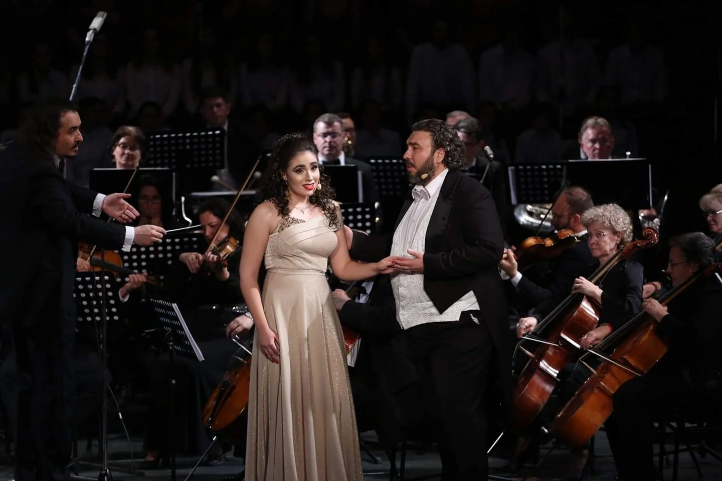 A woman in a champagne-colored gown and a man in a tuxedo exchange a touch during a live orchestral performance on stage.