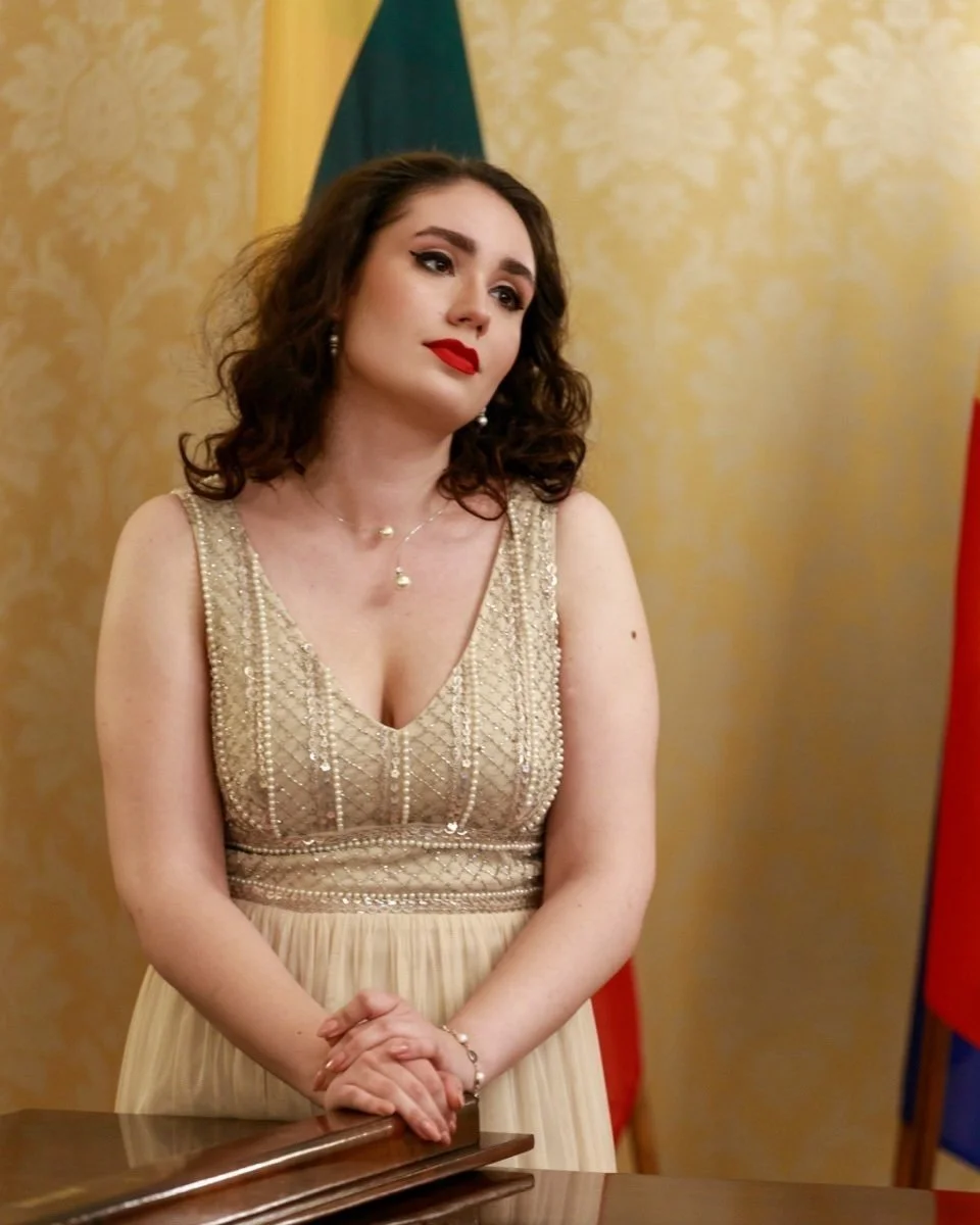 A young woman with dark curly hair, red lipstick, and makeup wearing a gold embellished dress, standing with her hands clasped on a table, in a formal setting with flags in the background.