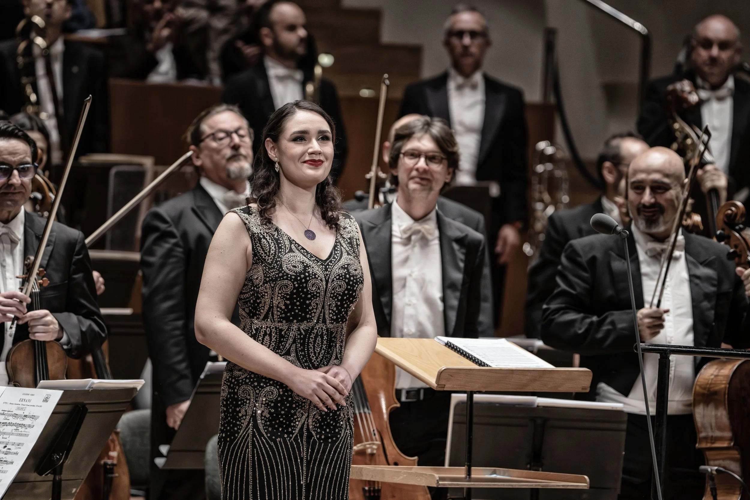 Orchestra conductor woman in a black and gold dress standing in front of an orchestra during a performance.