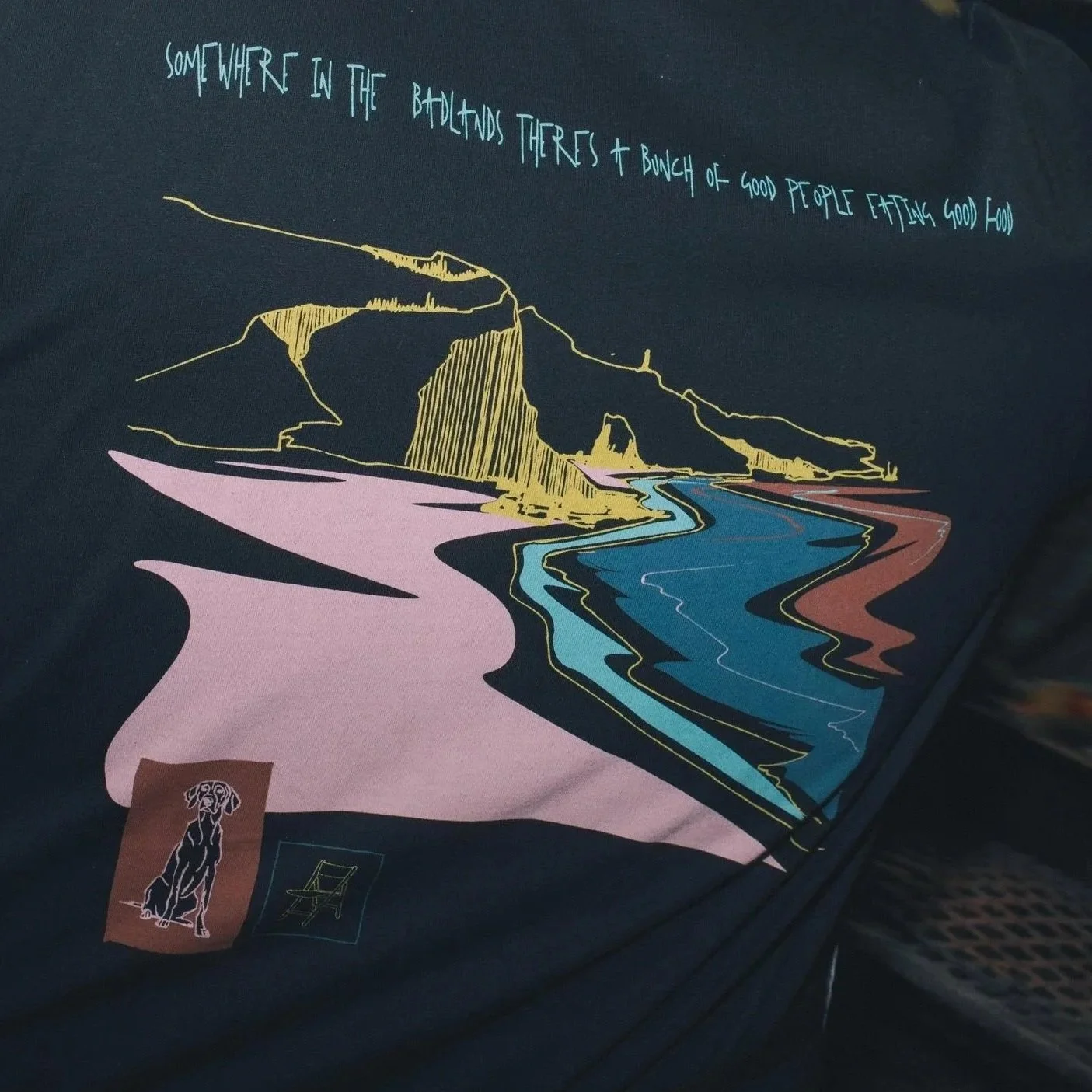 A graphic illustration of a landscape with a river flowing through a canyon, hills, and a trail. Text at the top reads, 'Somewhere in the backlands there's a bunch of good people eating good food.' There is a small logo of a dog at the bottom left corner.