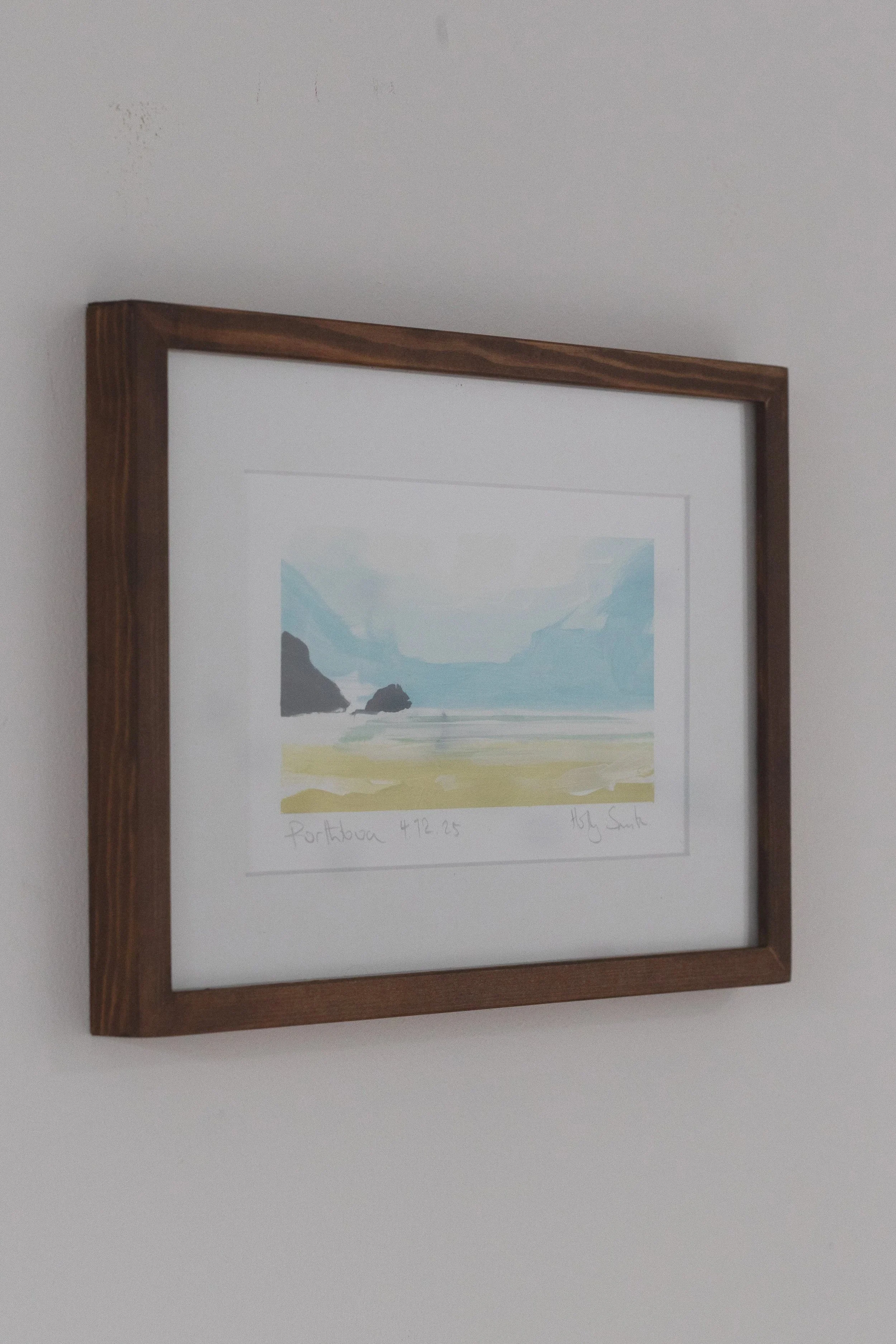 Framed watercolor painting of a coastal landscape, with cliffs, ocean, and sky, on a white wall.