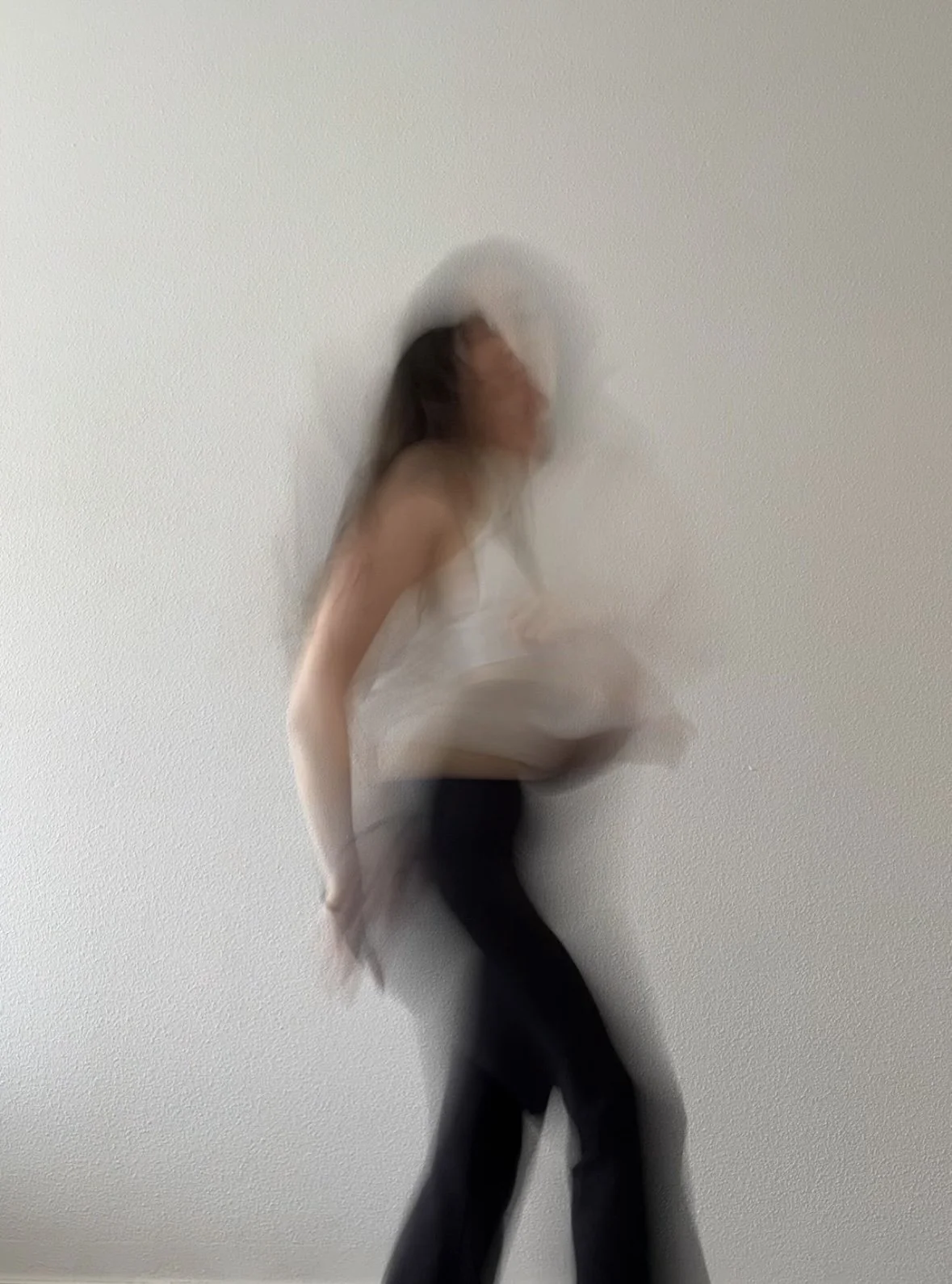 Blurry image of a woman standing against a plain white wall, holding a light-colored object, with abstract motion blur.