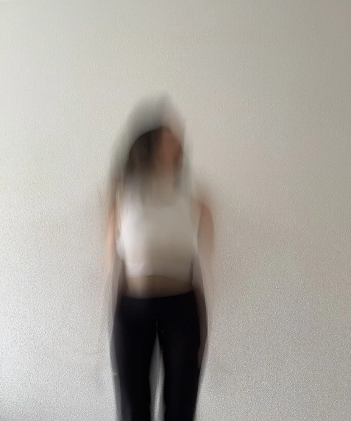 Blurred image of a woman standing in front of a plain wall, wearing a white top and black pants.