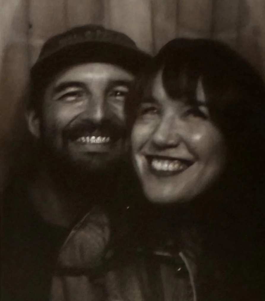 A black and white photo of a smiling man and woman posing closely together in front of a wooden background.