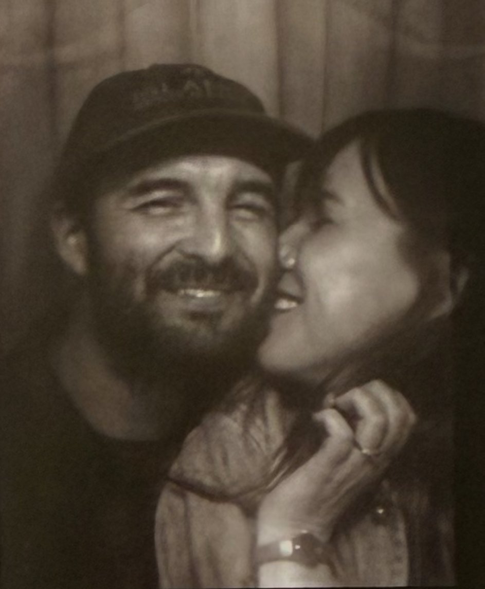 A woman is kissing a smiling man on the cheek in a close-up black-and-white photo with a wooden background.