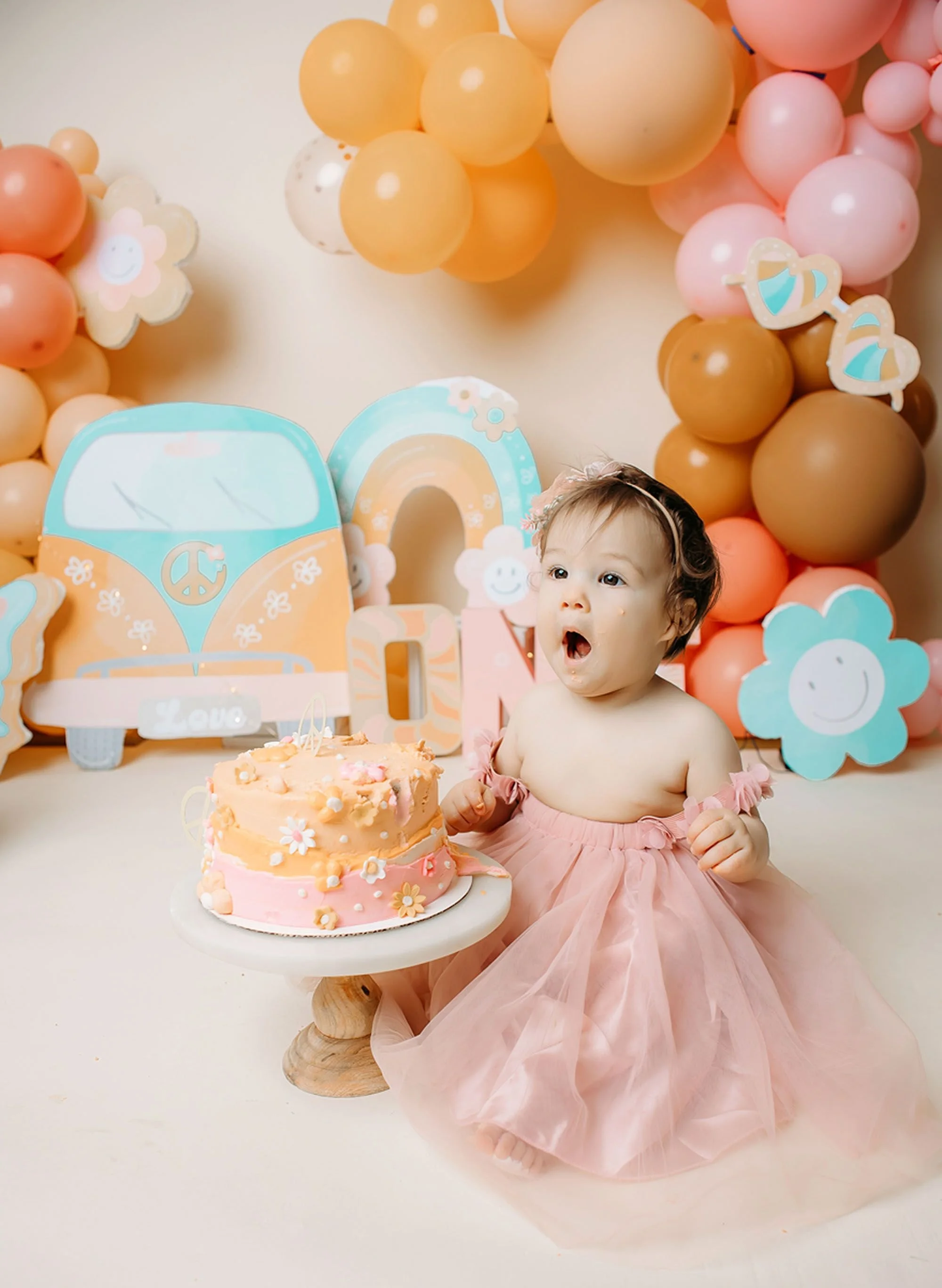 A little girl enjoys her first birthday cake beside a pastel pink cake, surrounded by groovy rainbow decorations, warm-toned balloons, and cheerful floral accents.