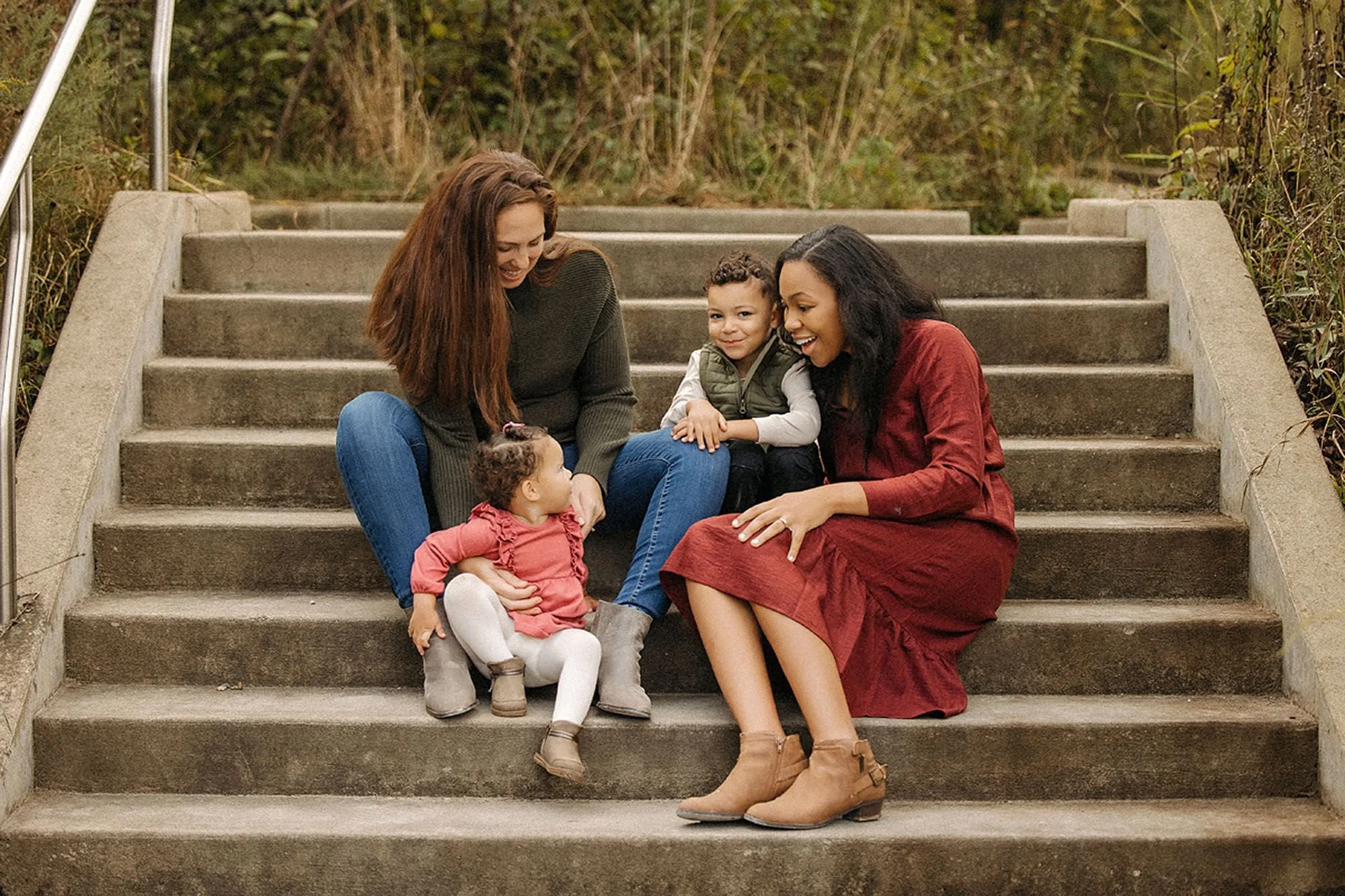 Two mothers and their toddlers sitting together on stone steps, smiling and chatting during a golden autumn family session at Virginia B. Fairbanks Art & Nature Park