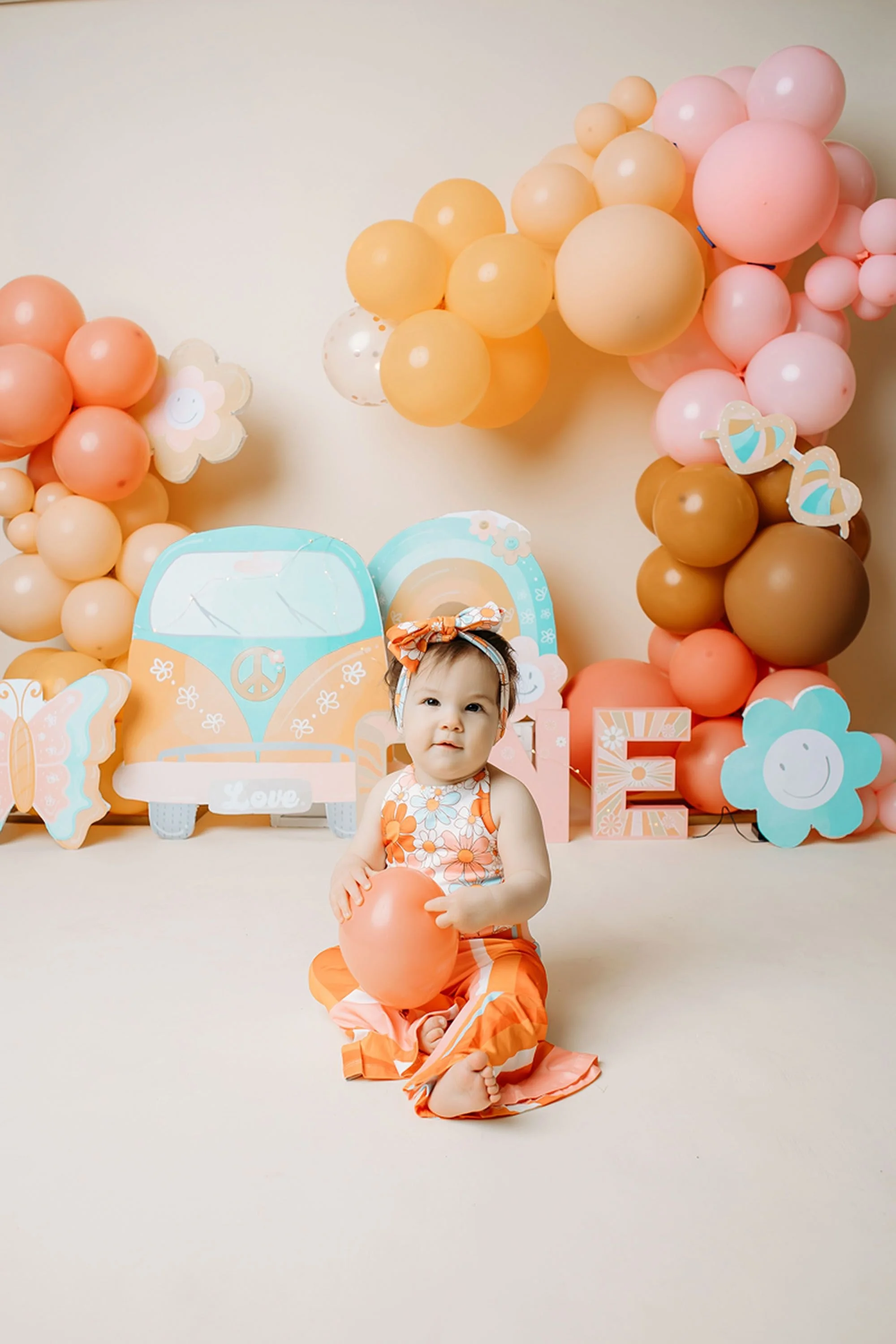A happy baby girl holds a peach balloon in a colorful Groovy One cake smash setup filled with pastel balloons, retro rainbow decor, flowers, and playful details.