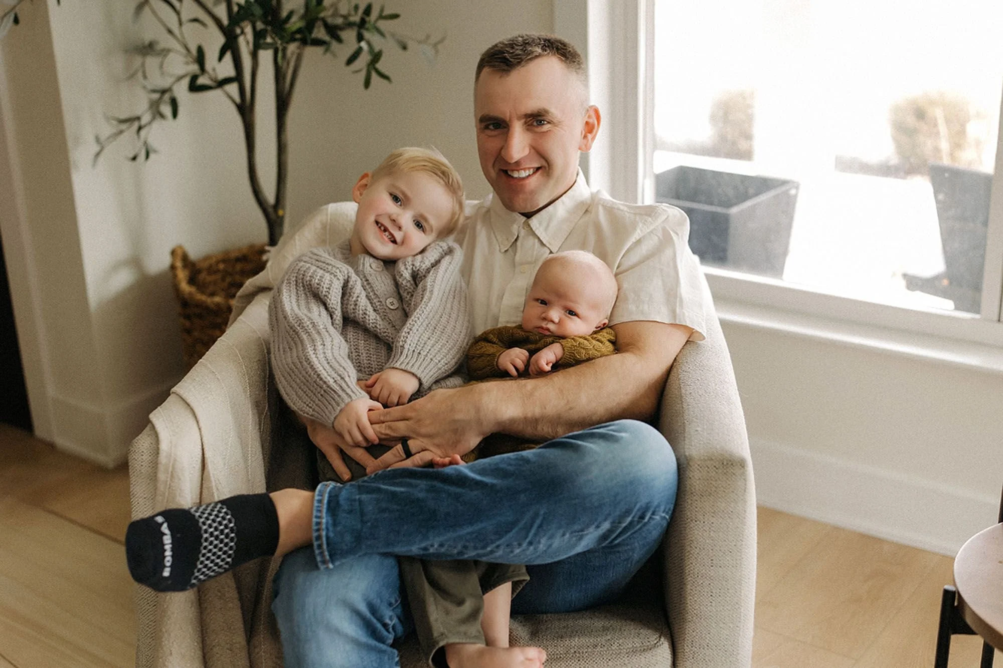 Newborn family portrait featuring a proud father holding his newborn baby and smiling toddler, and timeless in-home newborn photography in soft natural light