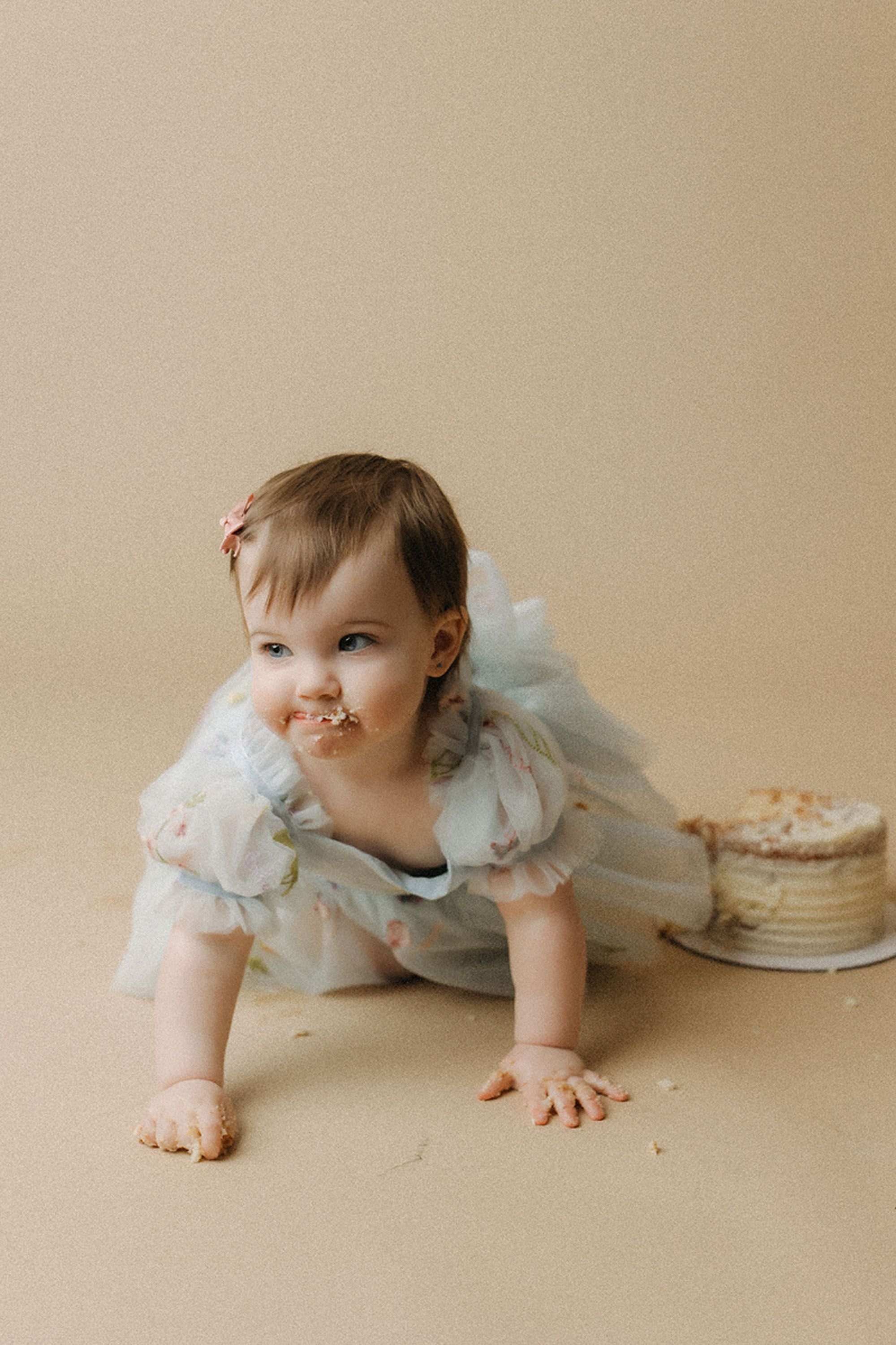Simple Cake Smash Photos featuring an adorable baby girl in a pastel blue dress crawling away from her birthday cake with a playful smile and cake crumbs around her mouth.