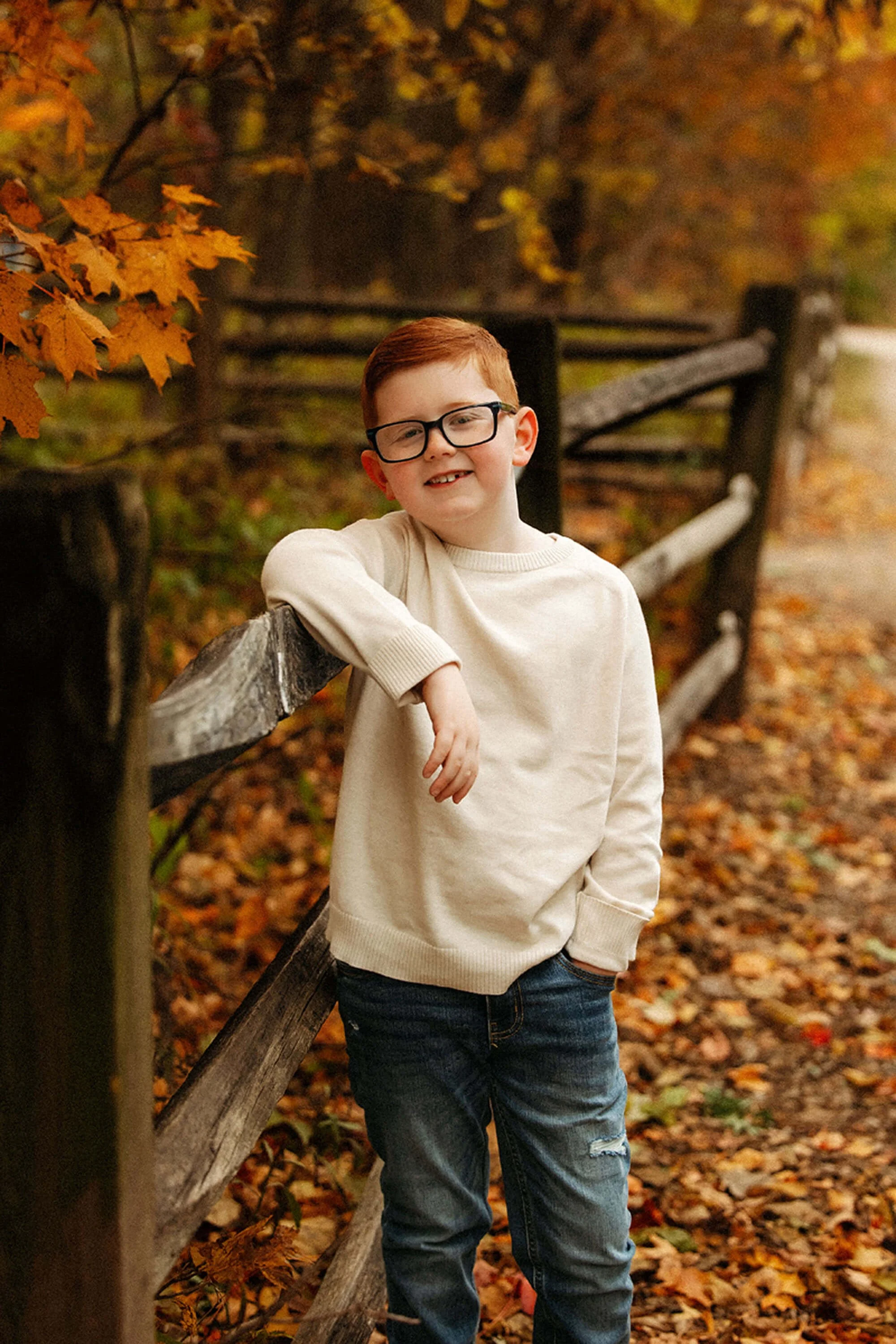 A Family Photographer captures a child's joyful personality as he smiles beside a rustic wooden fence, framed by rich autumn foliage.