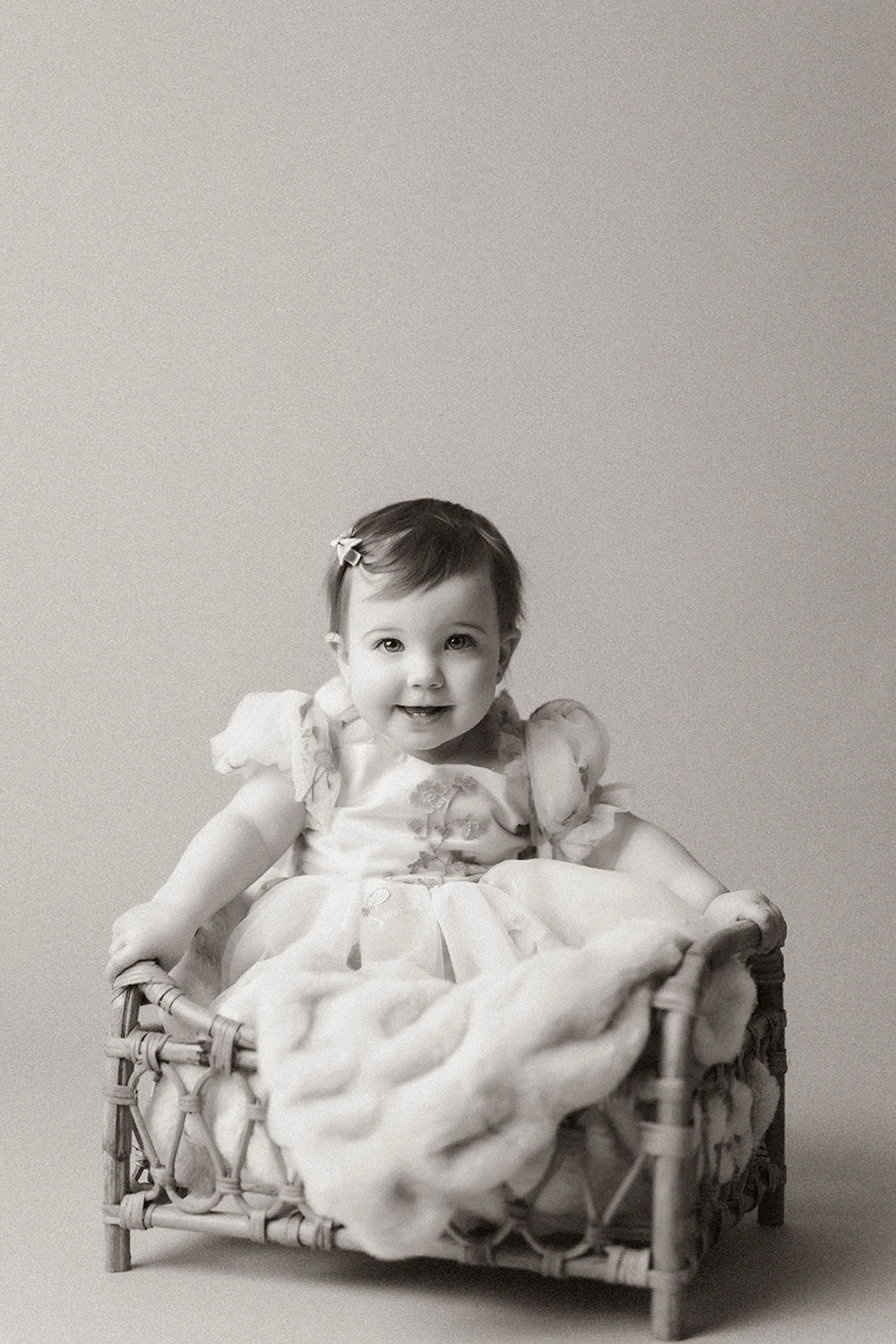 Simple Cake Smash Photos of a smiling baby girl sitting in a rustic wooden chair, wearing a delicate white dress and looking sweetly at the camera.