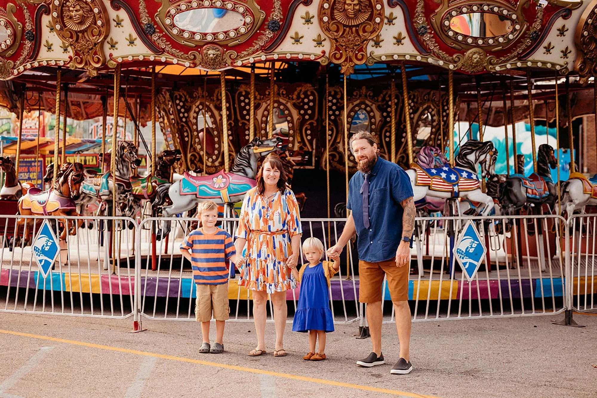 Family stands together, smiling in front of a brightly lit carousel during a carnival photo session in Indiana