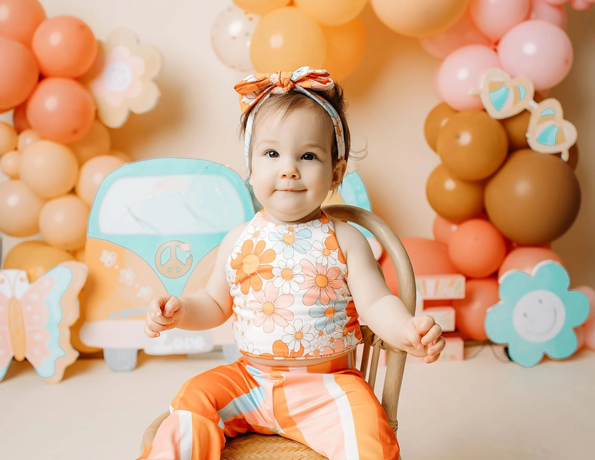 Smiling baby girl in a retro floral outfit posing for Groovy One cake smash photos in Indianapolis with a pastel VW bus backdrop and colorful balloon display