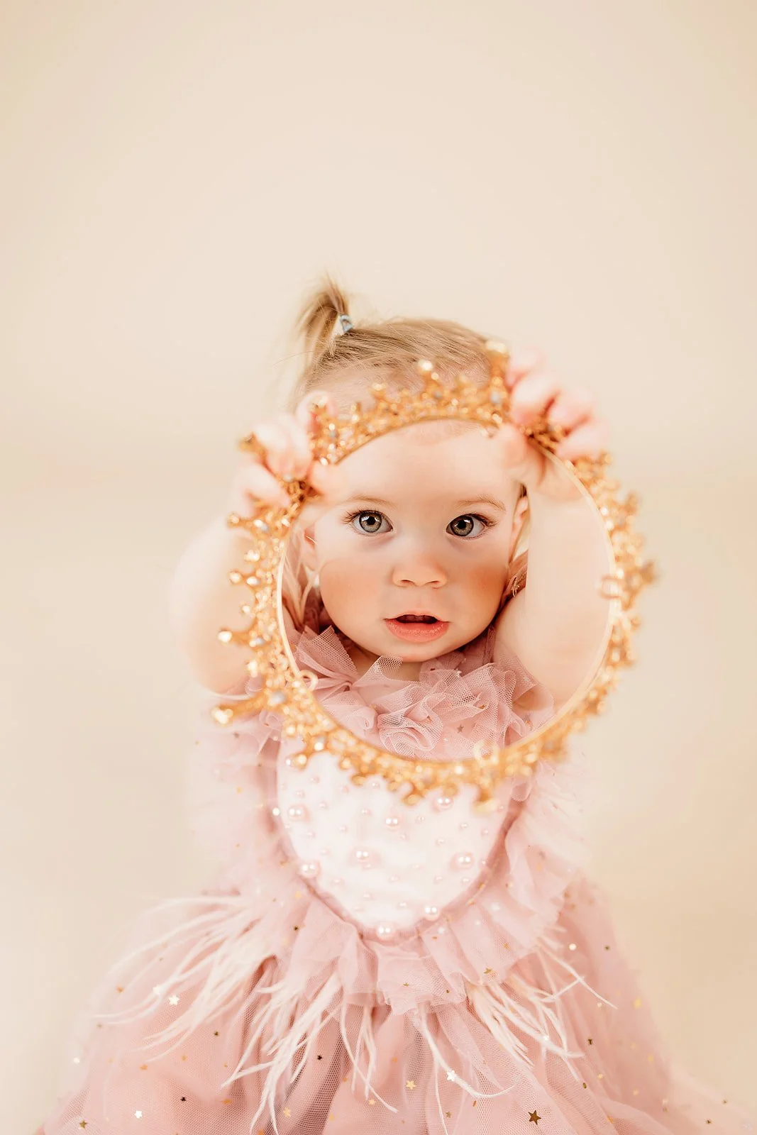 A young girl with blonde hair in a ponytail wearing a pink dress and a glittery pink tiara, looking into a decorative round mirror with a gold frame, with her arms raised holding the mirror in front of her face.