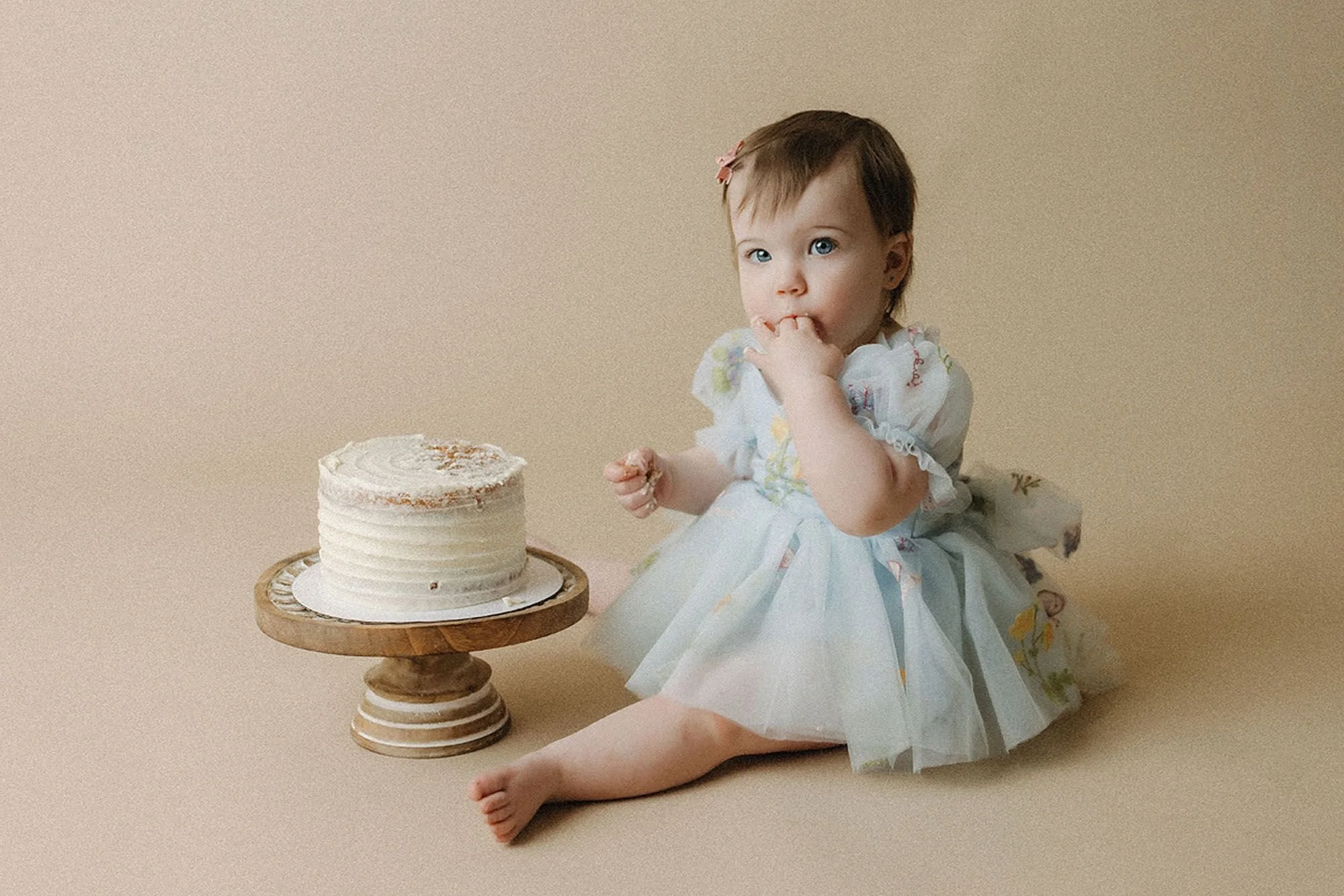 Baby girl in a light blue floral dress sitting beside an untouched white buttercream cake on a wooden stand, tasting frosting from her fingers, simple cake smash photography setup
