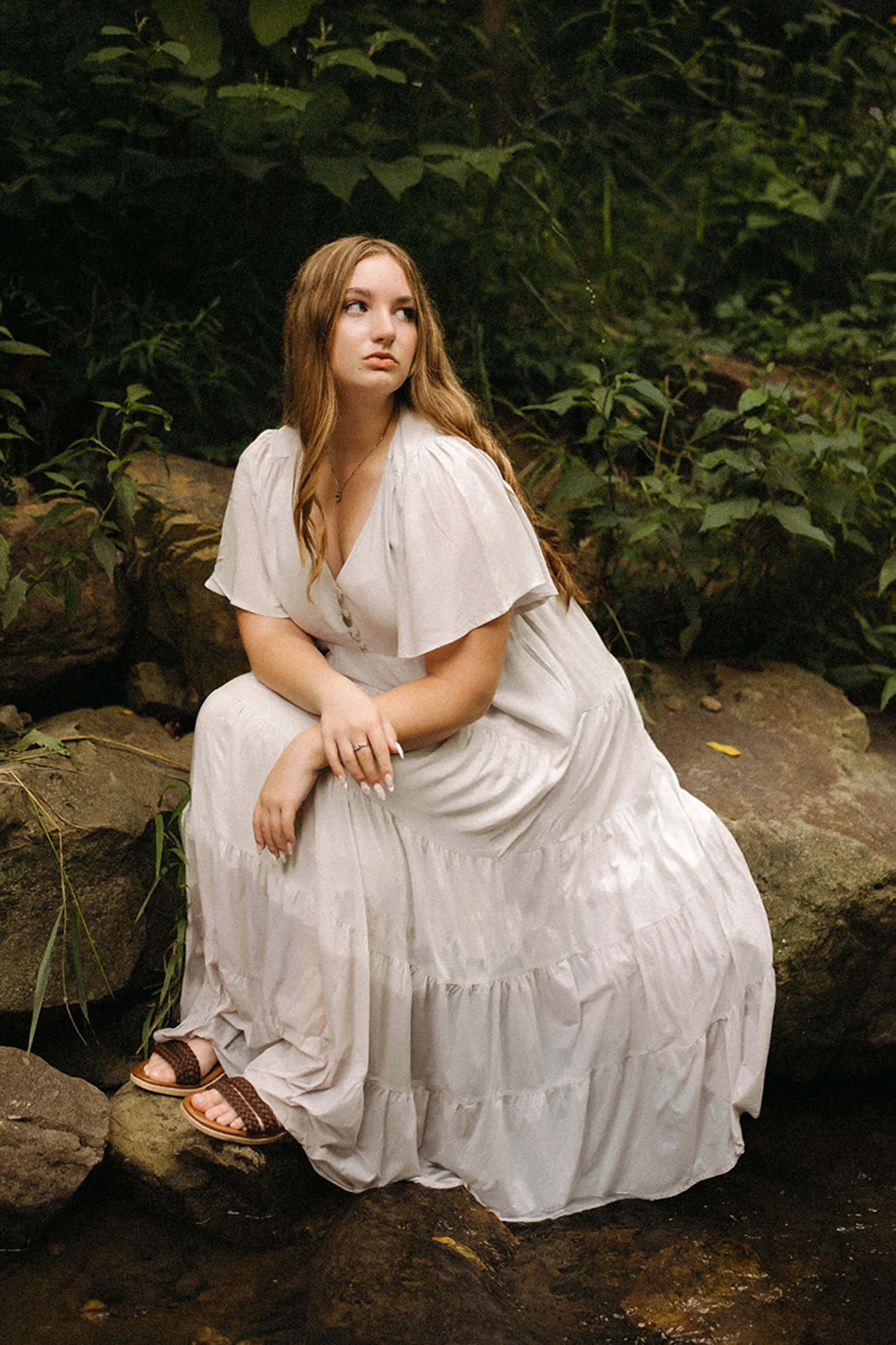Senior girl in a flowing white dress posing on natural rocks surrounded by lush greenery during her outdoor senior portrait session