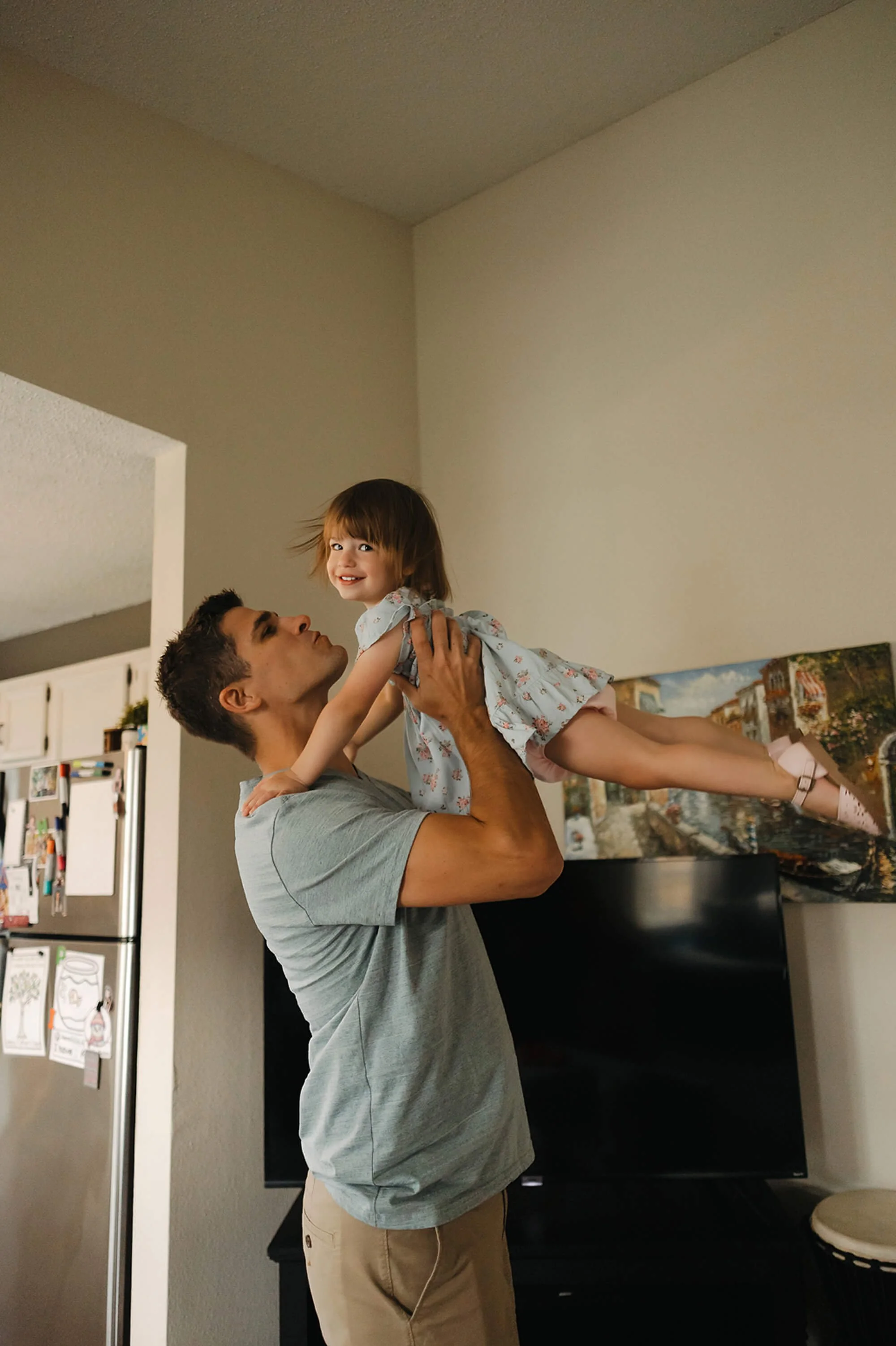 Father lifts smiling toddler daughter in the air during a playful in-home family photography session in Fishers, Indiana, capturing a joyful candid moment