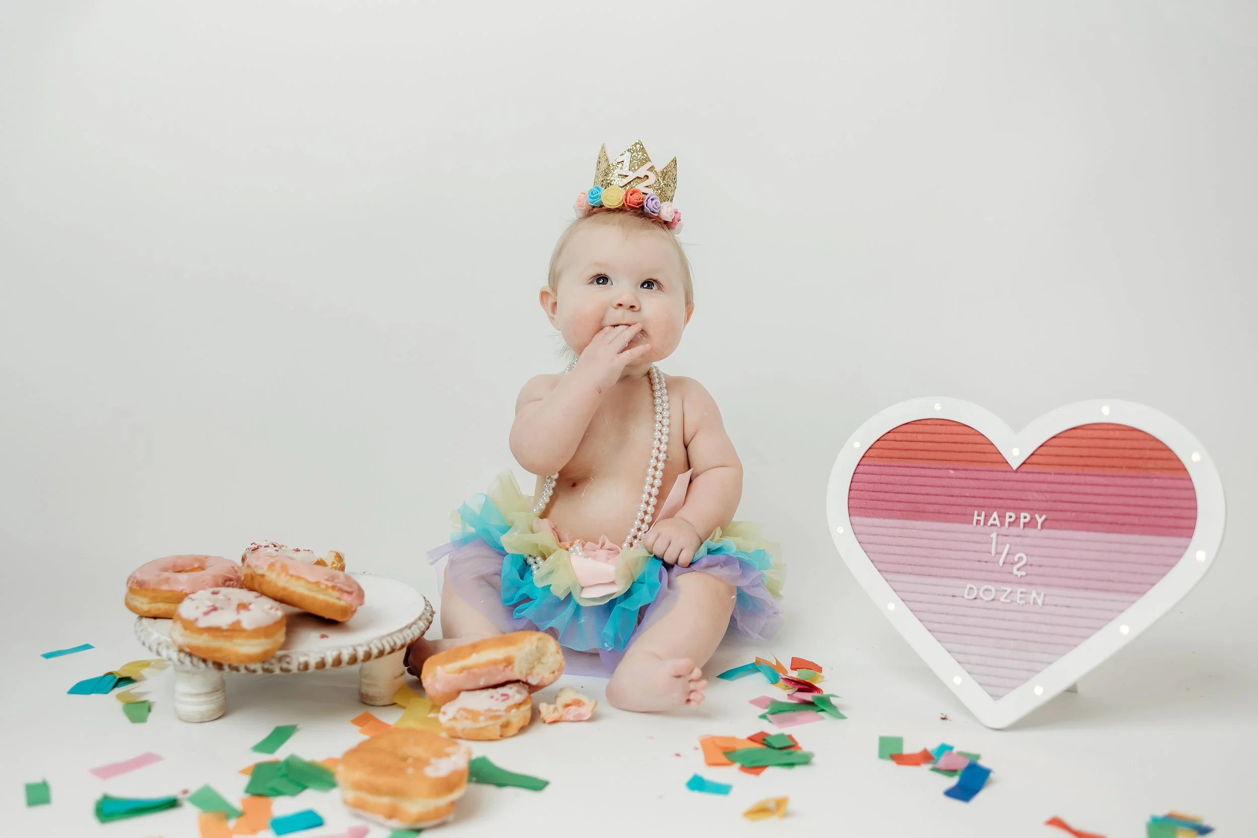 baby girl dressed in a colorful tutu and tiara sits in front of a heart-shaped cake, marking her 6-month milestone